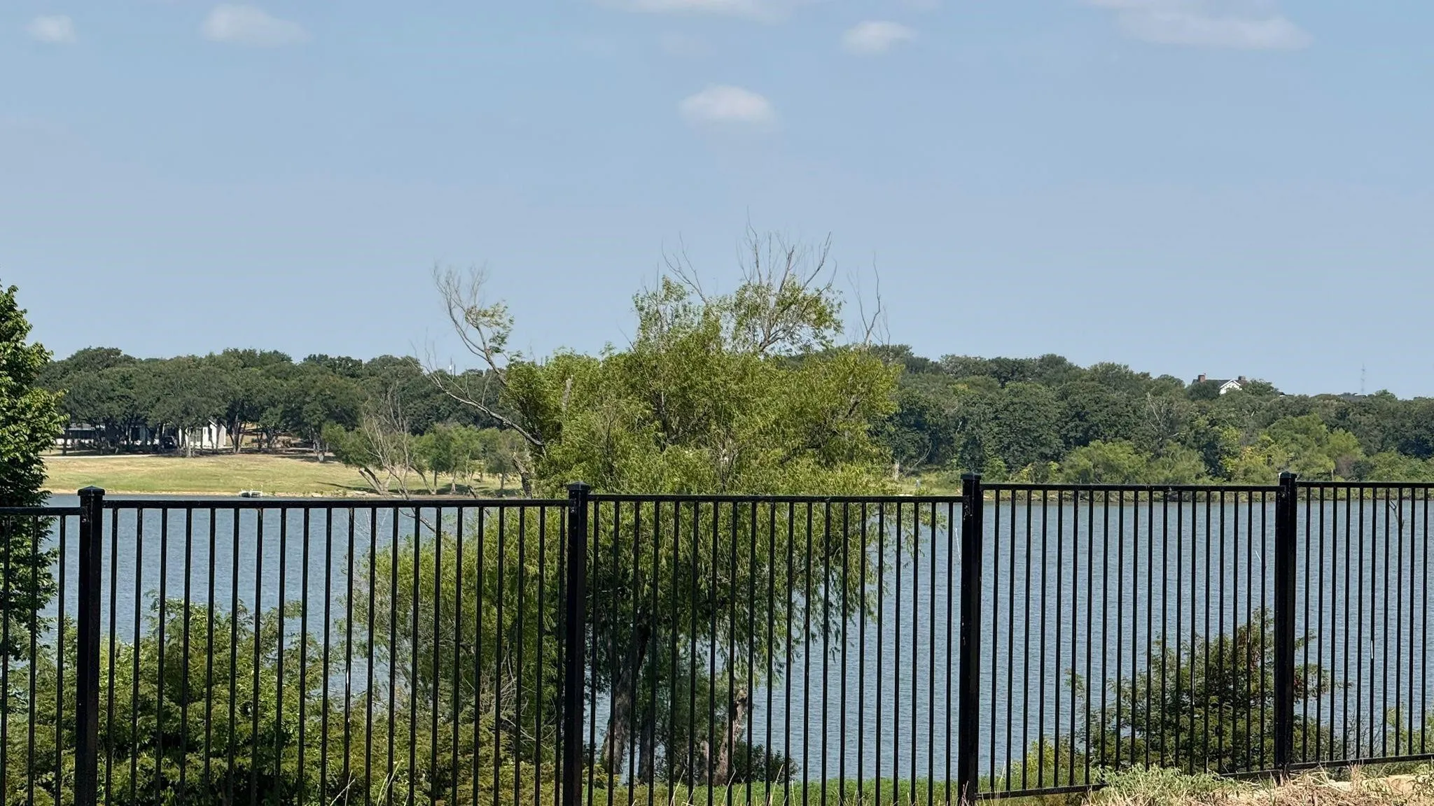 Waterfront property featuring a black metal fence, mature trees, and a distant treeline