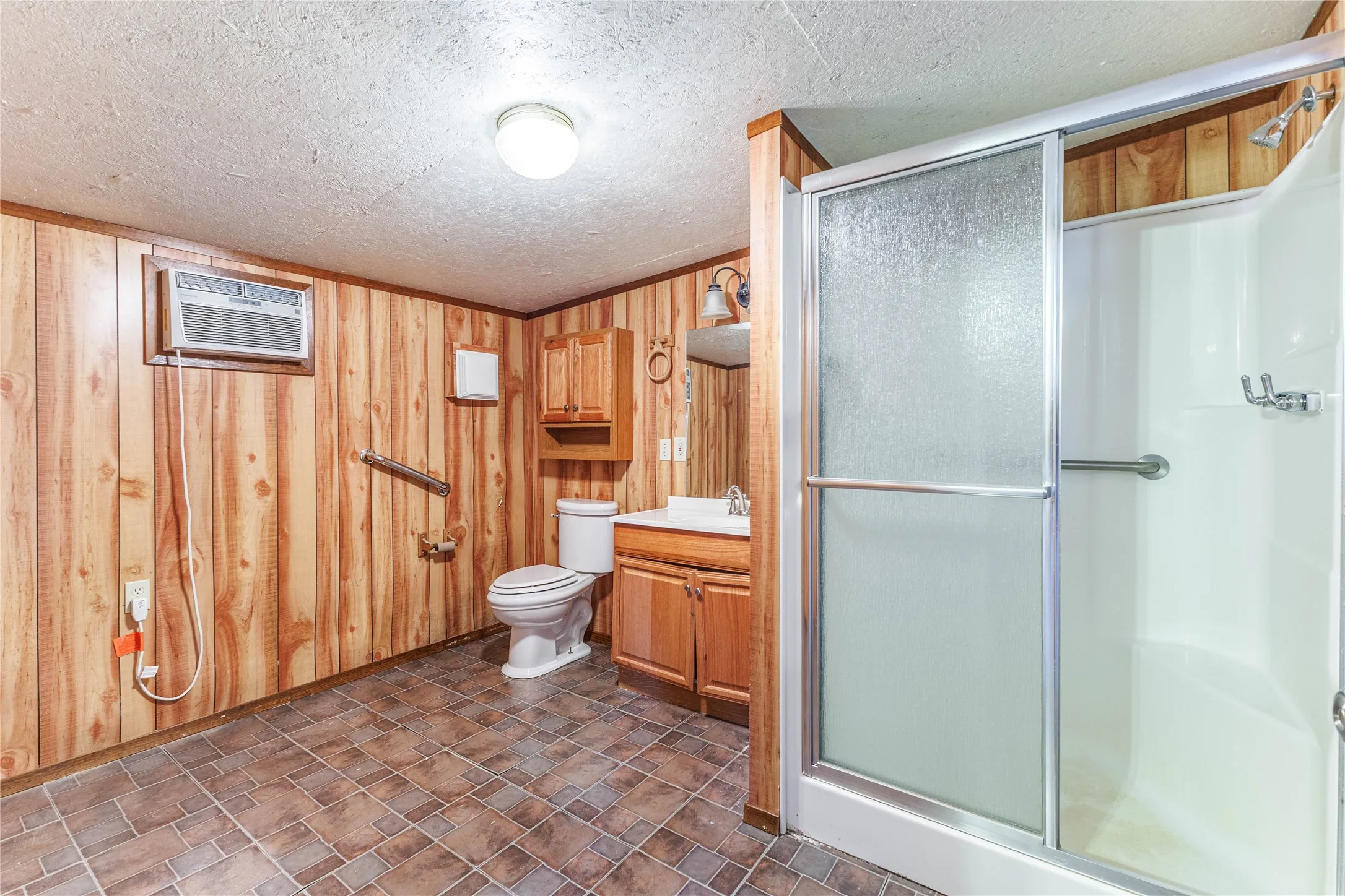 Shower enclosure with sliding frosted glass door, wall-mounted air conditioning unit, wood-paneled walls, and tiled flooring