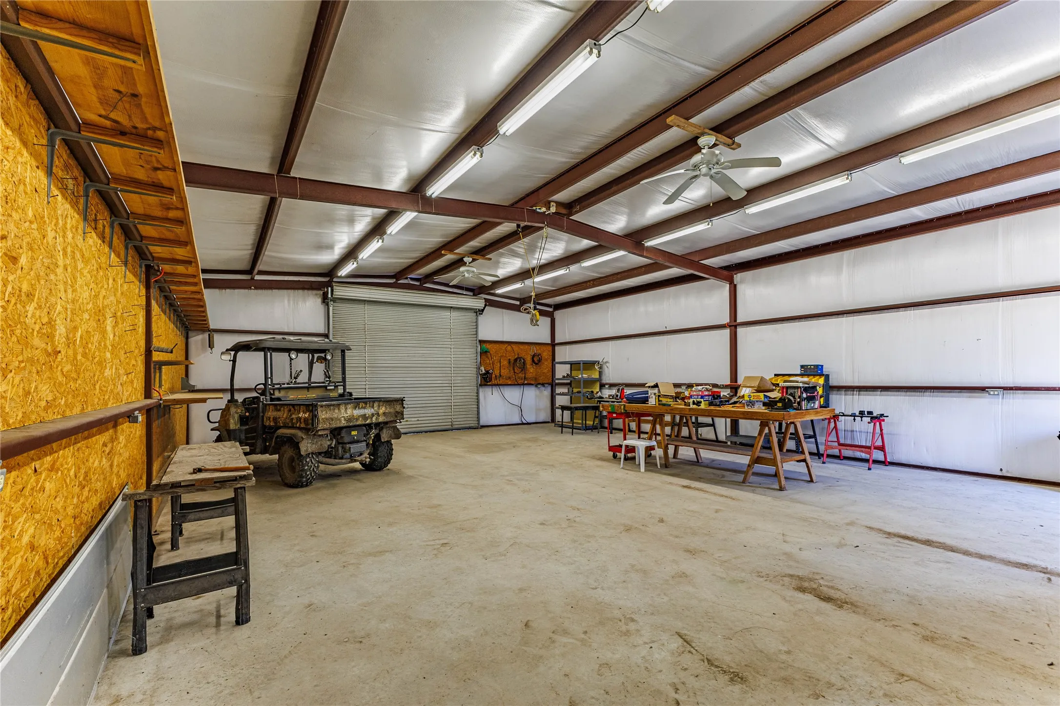 Spacious outbuilding featuring a concrete floor, exposed metal beam ceiling, flush-mounted fluorescent lighting, a roll-up door, and ceiling fans