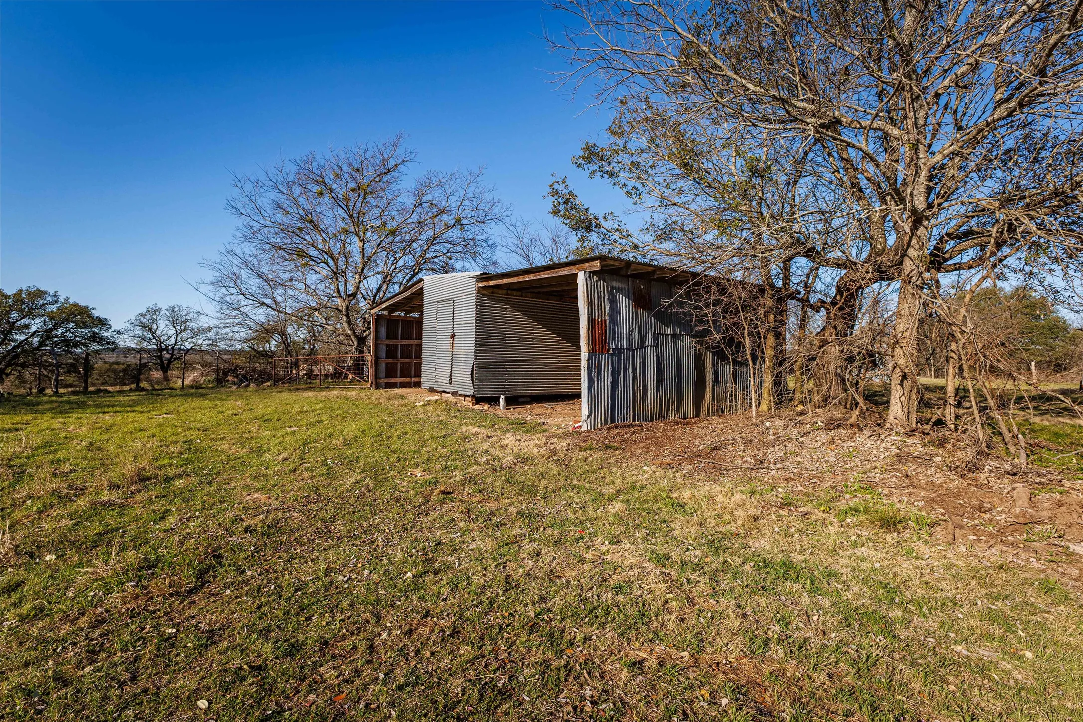 Metal-clad structure with an open-sided shed area and an enclosed storage space, set on a grassy expanse with mature trees