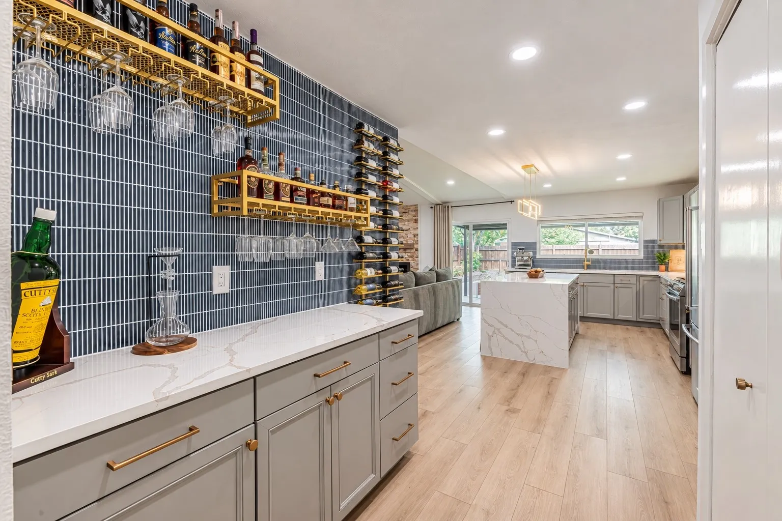Vaulted ceilings with recessed lighting, light wood-finish flooring, kitchen island with a waterfall countertop, gray cabinetry with brass hardware, and a blue tile backsplash