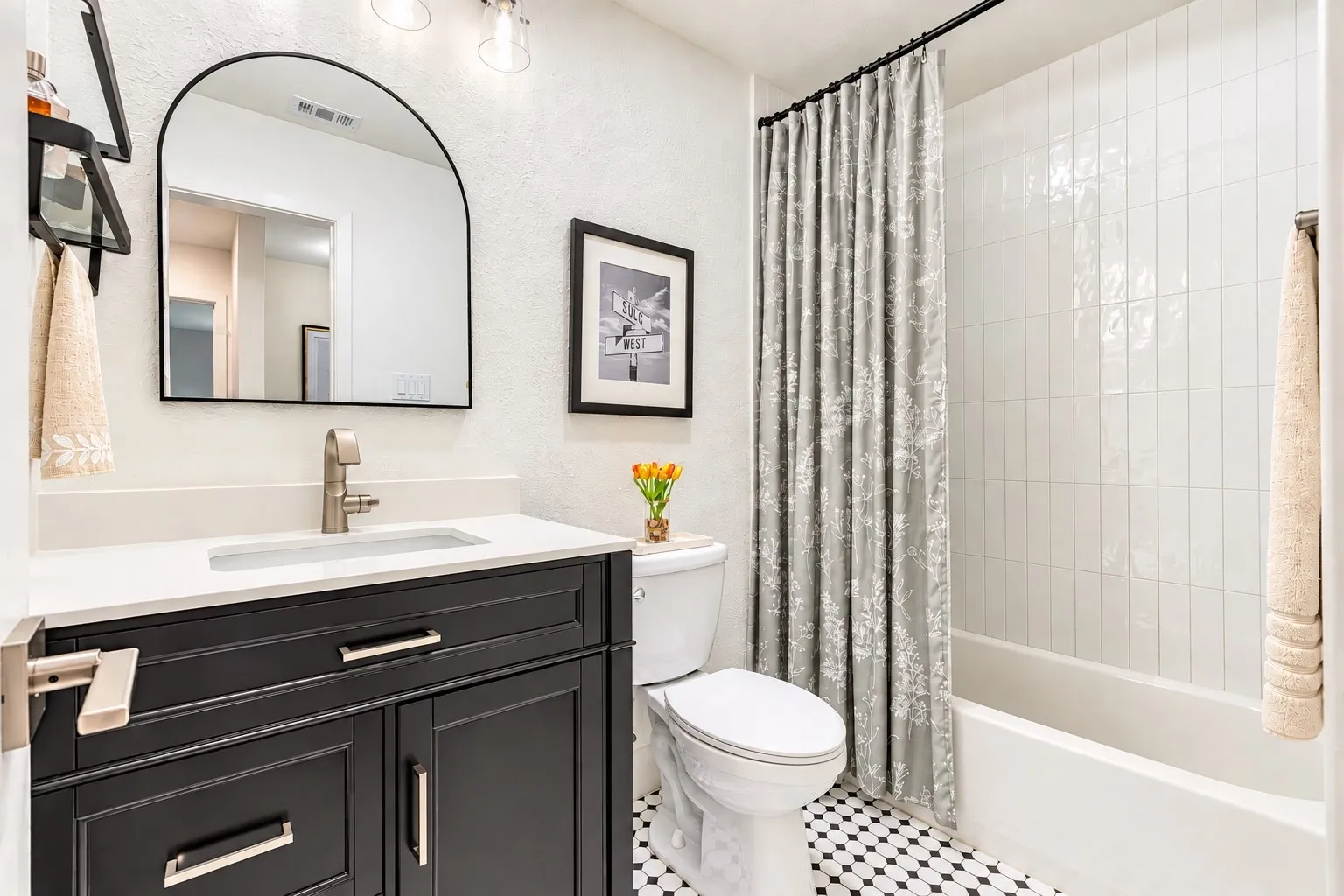 Black vanity with brushed nickel hardware, white countertop, arched mirror, tiled shower surround, and black and white floor tiling