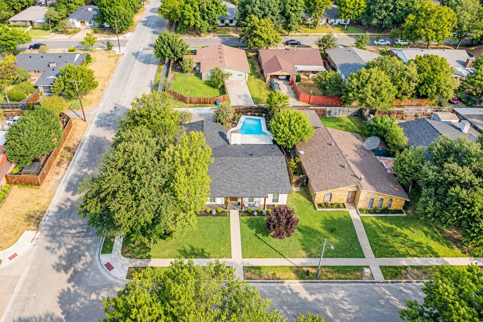 Aerial perspective showcasing a single-story residence with a dark shingle roof, a rectangular swimming pool, and mature trees
