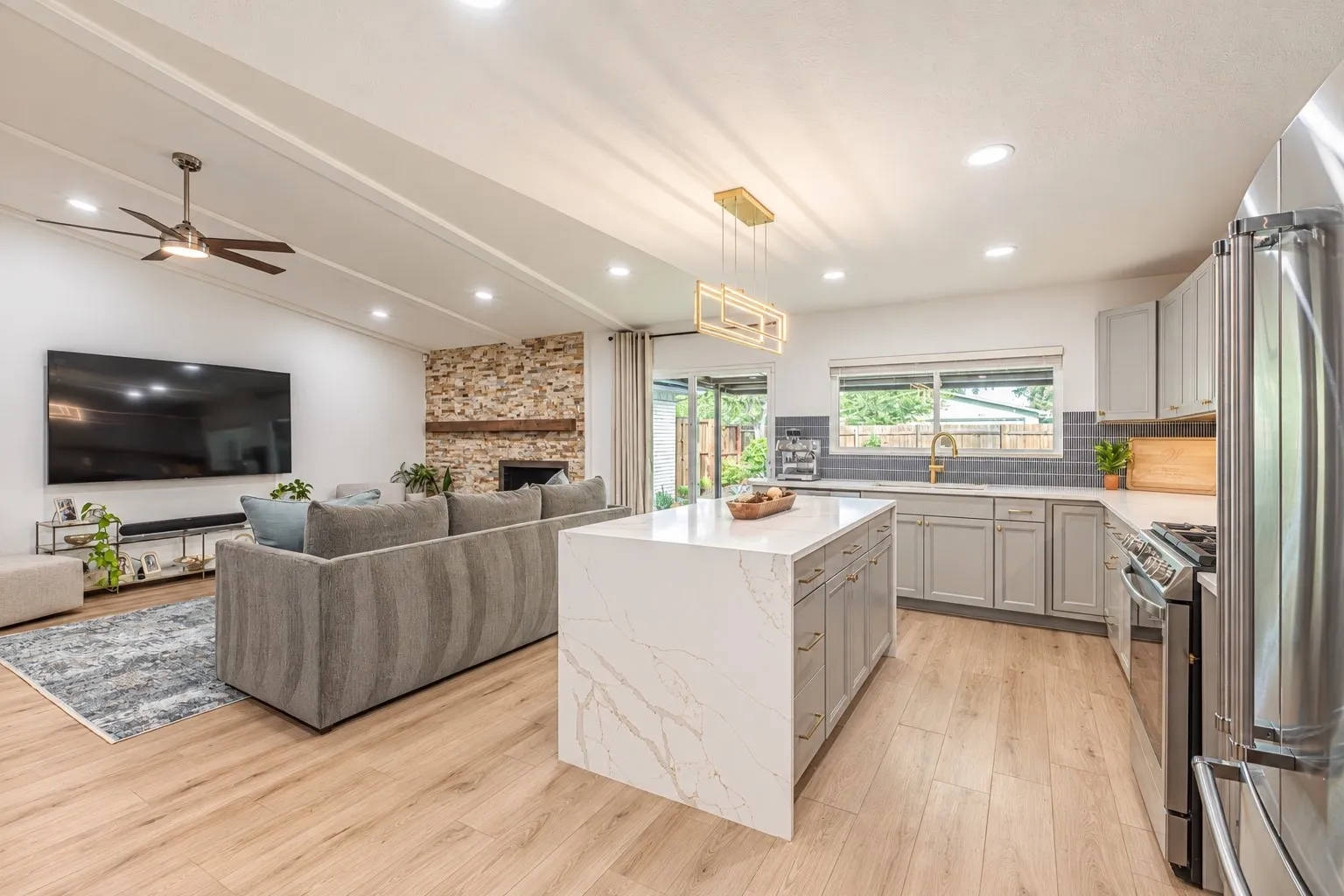 Open-concept living space featuring vaulted ceilings, wood-finish flooring, a stone-clad fireplace with wood mantel, a kitchen island with stone countertop, and a kitchen with stainless steel appliances