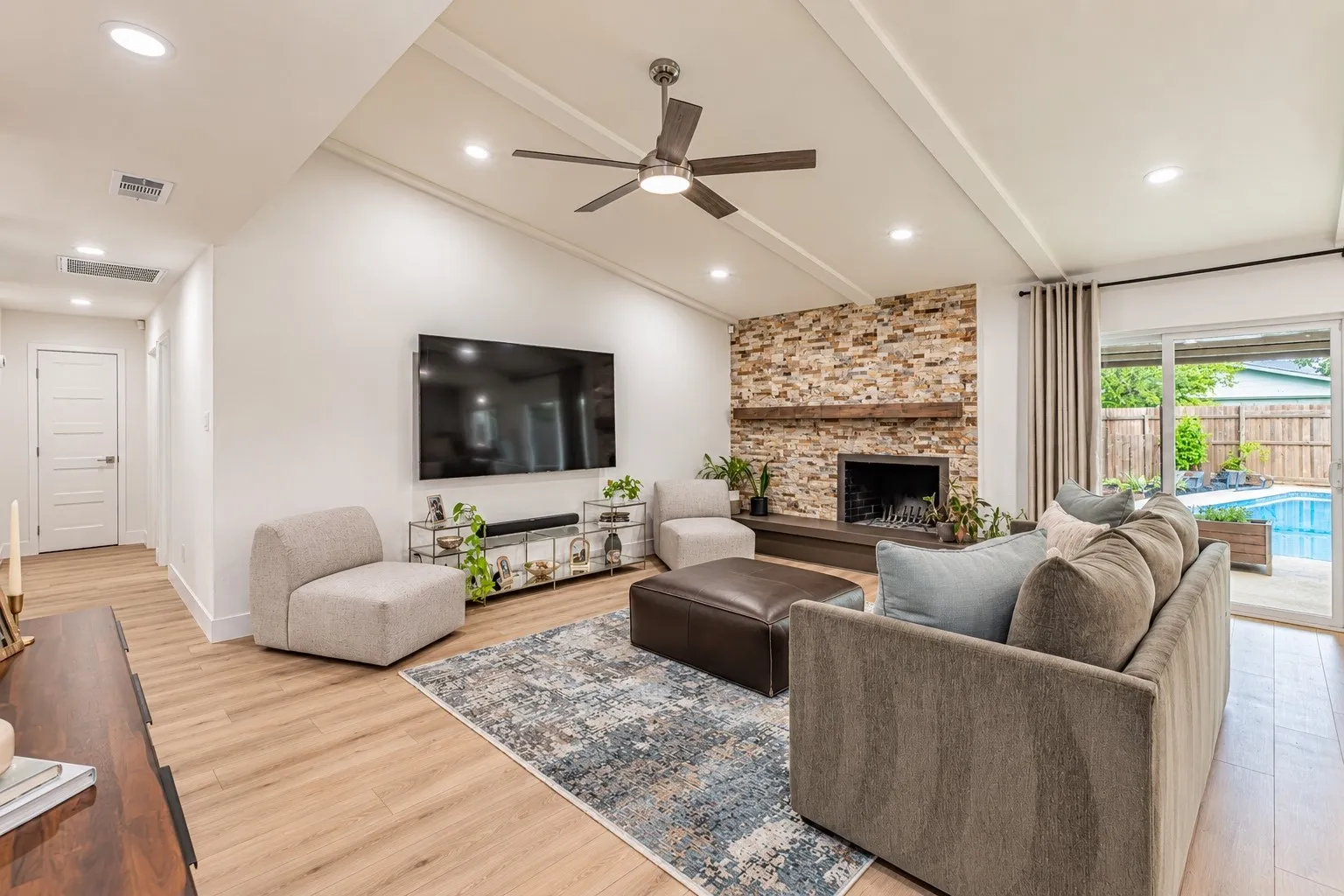 Vaulted ceiling with exposed beams, recessed lighting, wood-finish flooring, stone-clad fireplace, and sliding glass door to an outdoor pool
