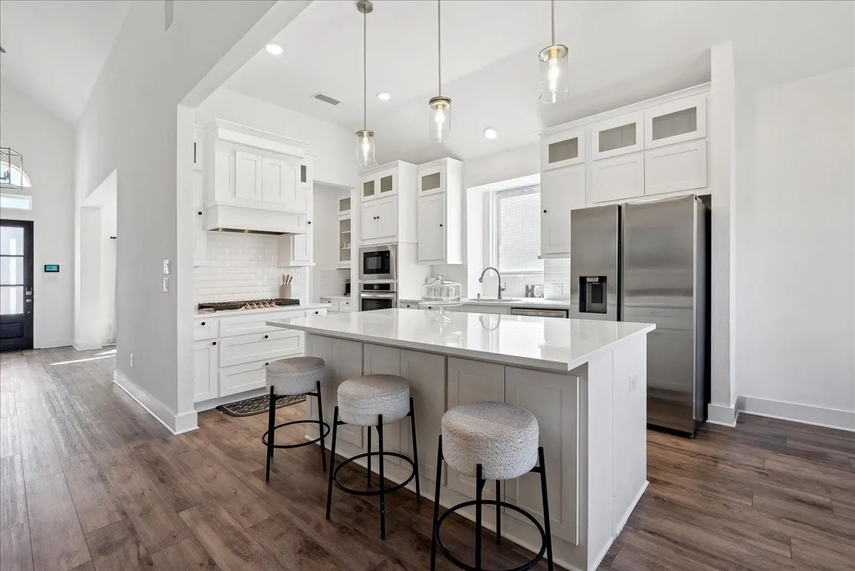 White cabinetry and quartz countertops define this kitchen, featuring an island with pendant lighting, wood-finish flooring, and stainless steel appliances