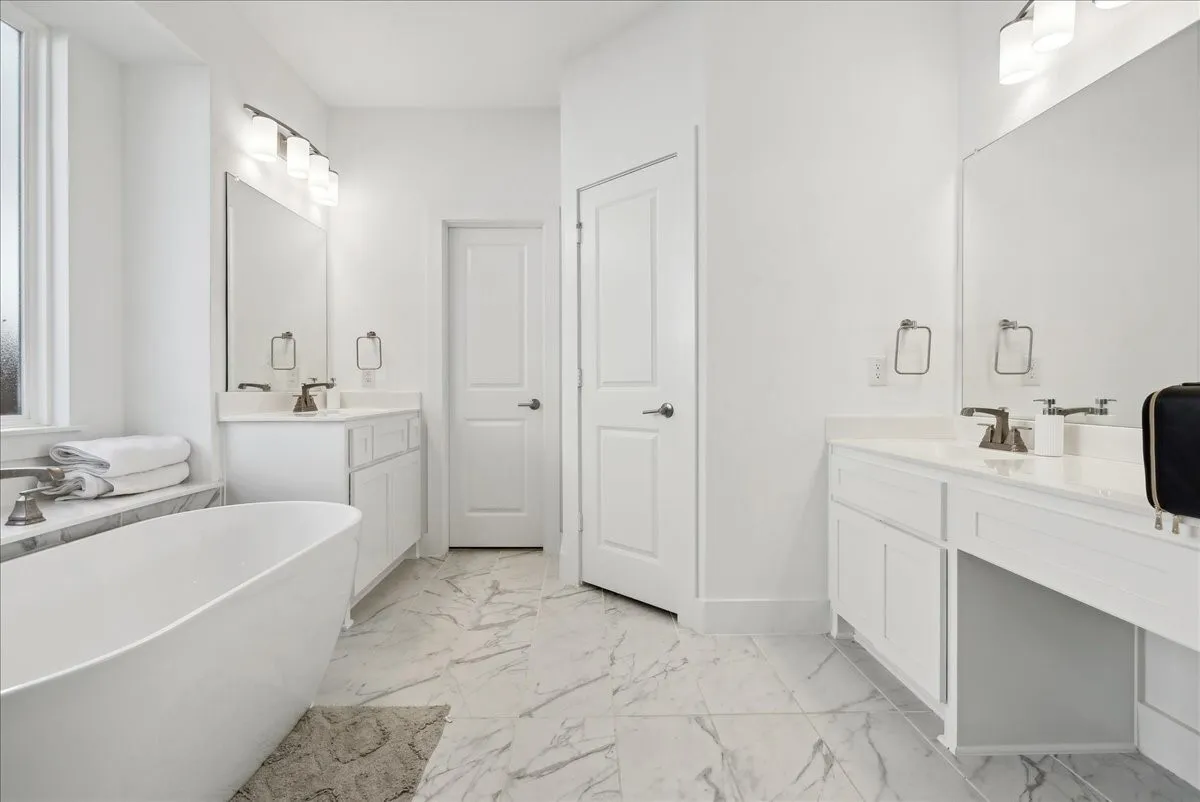 Freestanding soaking tub, dual vanity with undermount sinks, white cabinetry, and large format tile flooring