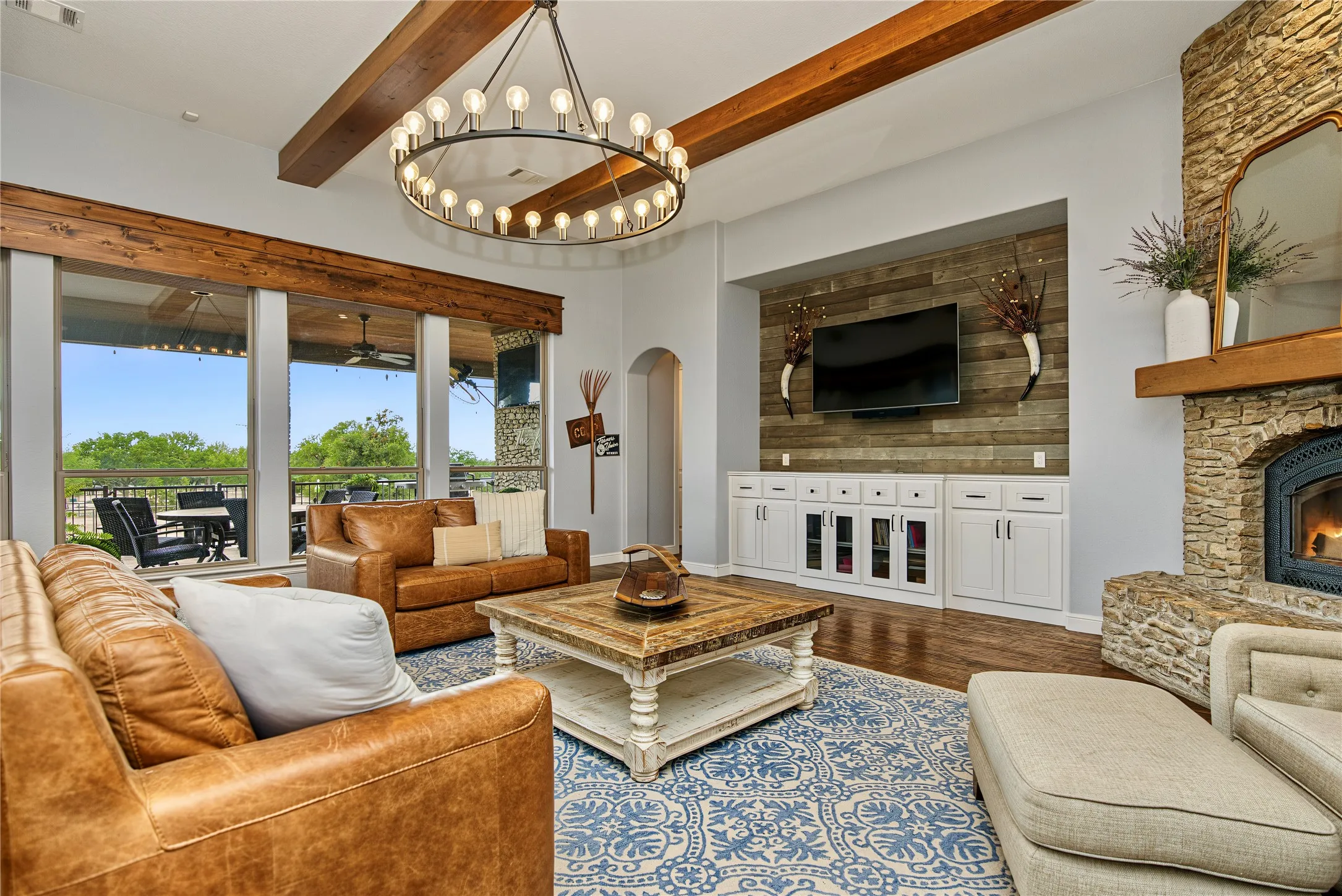 Living area featuring a stone fireplace, wood-finish flooring, exposed wood ceiling beams, and a large multi-pane window overlooking an outdoor patio