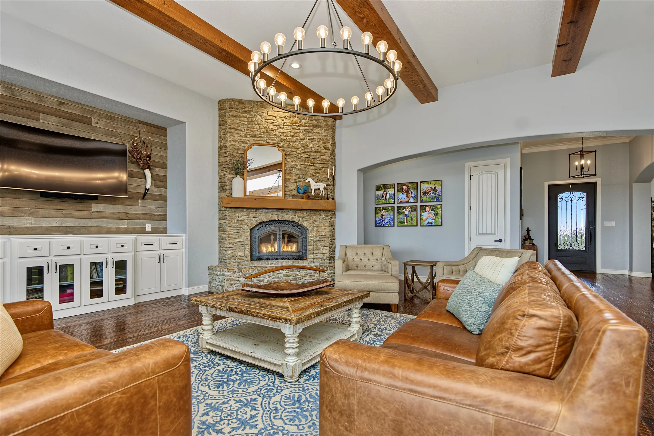Vaulted ceiling with wood beams, stone-clad fireplace, built-in media cabinetry with wood-finish accent wall, dark wood-finish flooring, and arched entryway