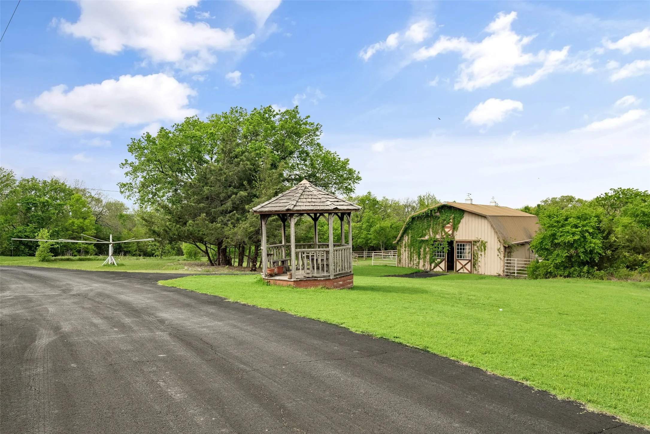 Asphalt drive, wooden gazebo with shingle roof, expansive green lawn, mature trees, and a barn structure