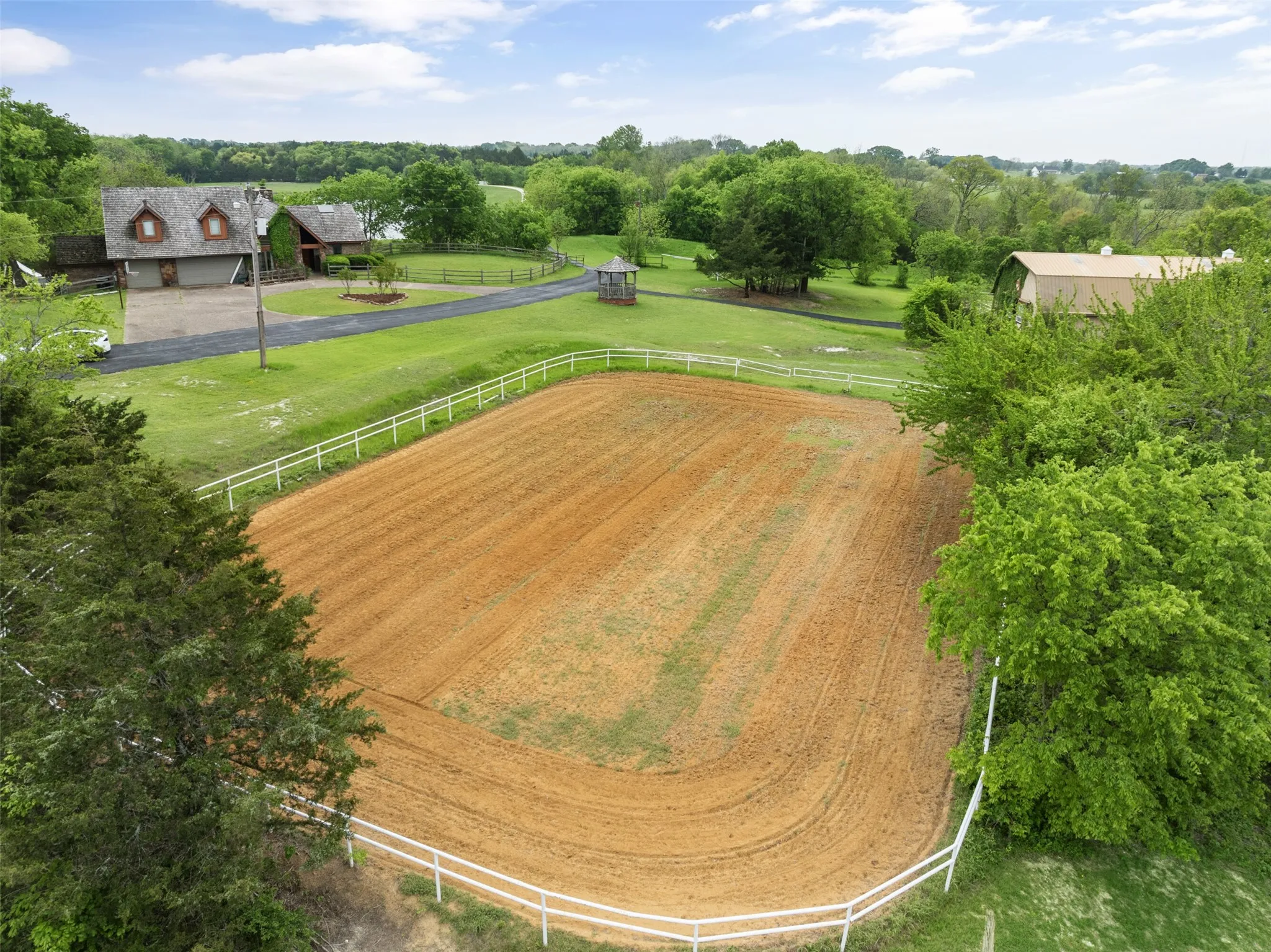 Spacious equestrian arena with white fencing, bordered by mature trees and expansive green grounds