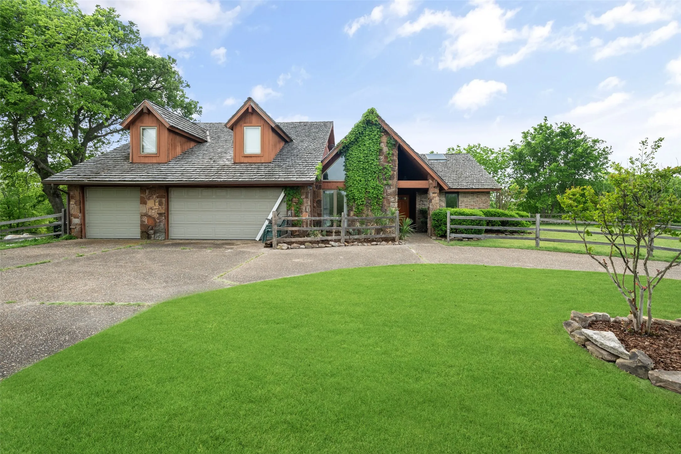 Stone and wood-sided exterior featuring an attached two-car garage, dormer windows, and a fenced front yard with a paved driveway