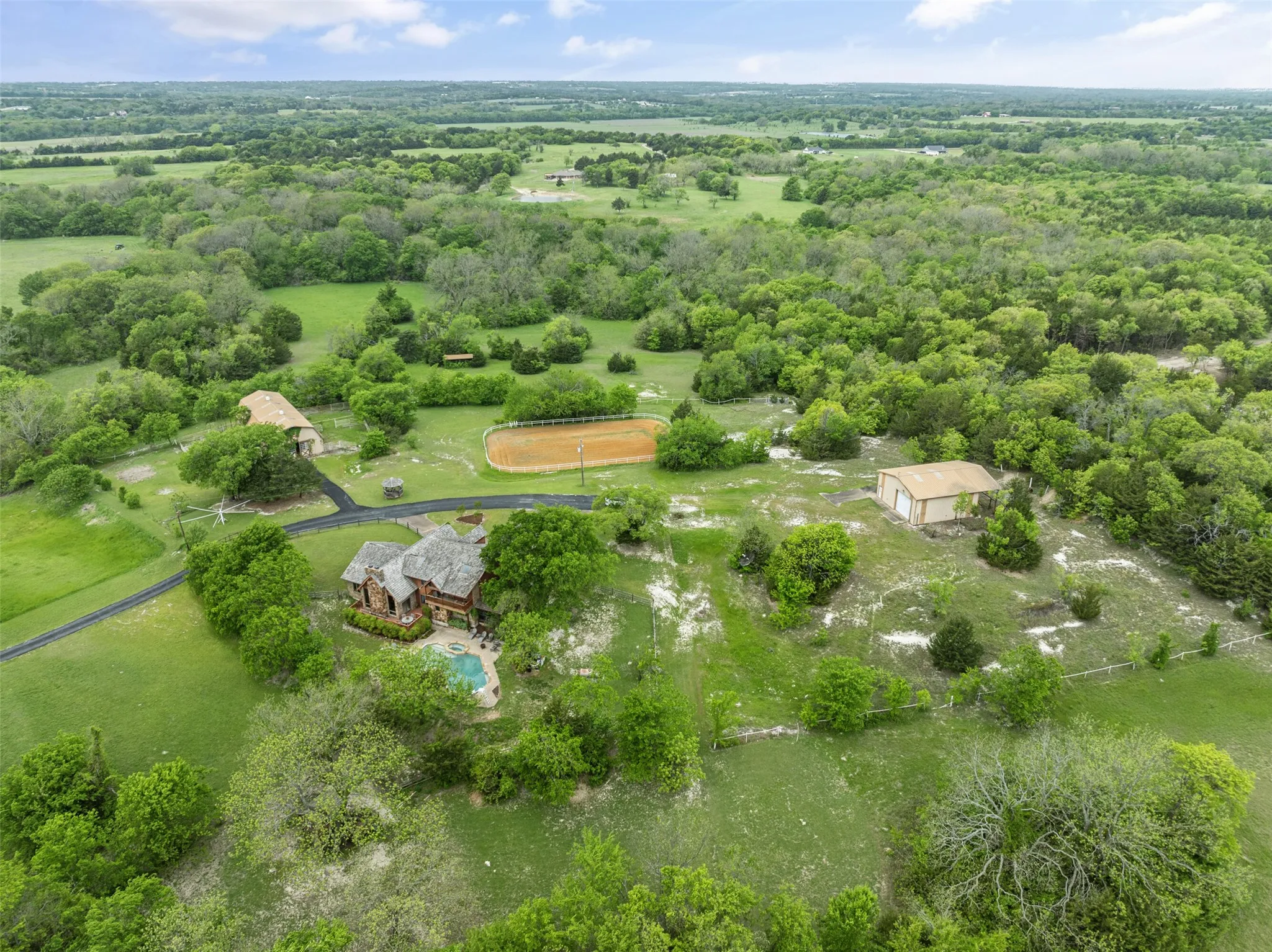 Sprawling acreage featuring a main residence with a swimming pool, a separate barn structure, an equestrian arena, and an additional outbuilding, all surrounded by extensive natural landscaping