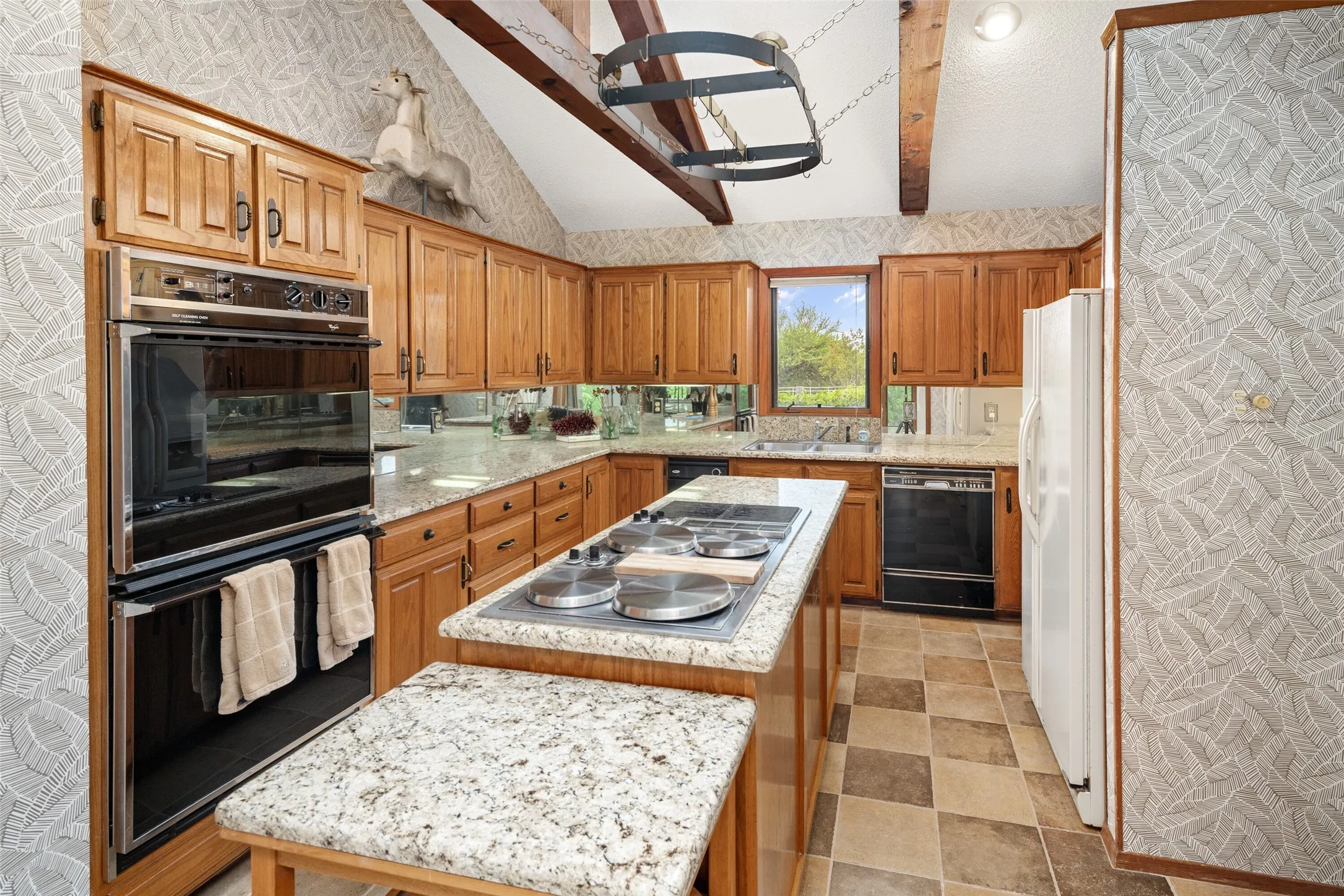 Vaulted ceiling with exposed wood beams, wood cabinetry, built-in double oven, island with cooktop, and patterned tile flooring