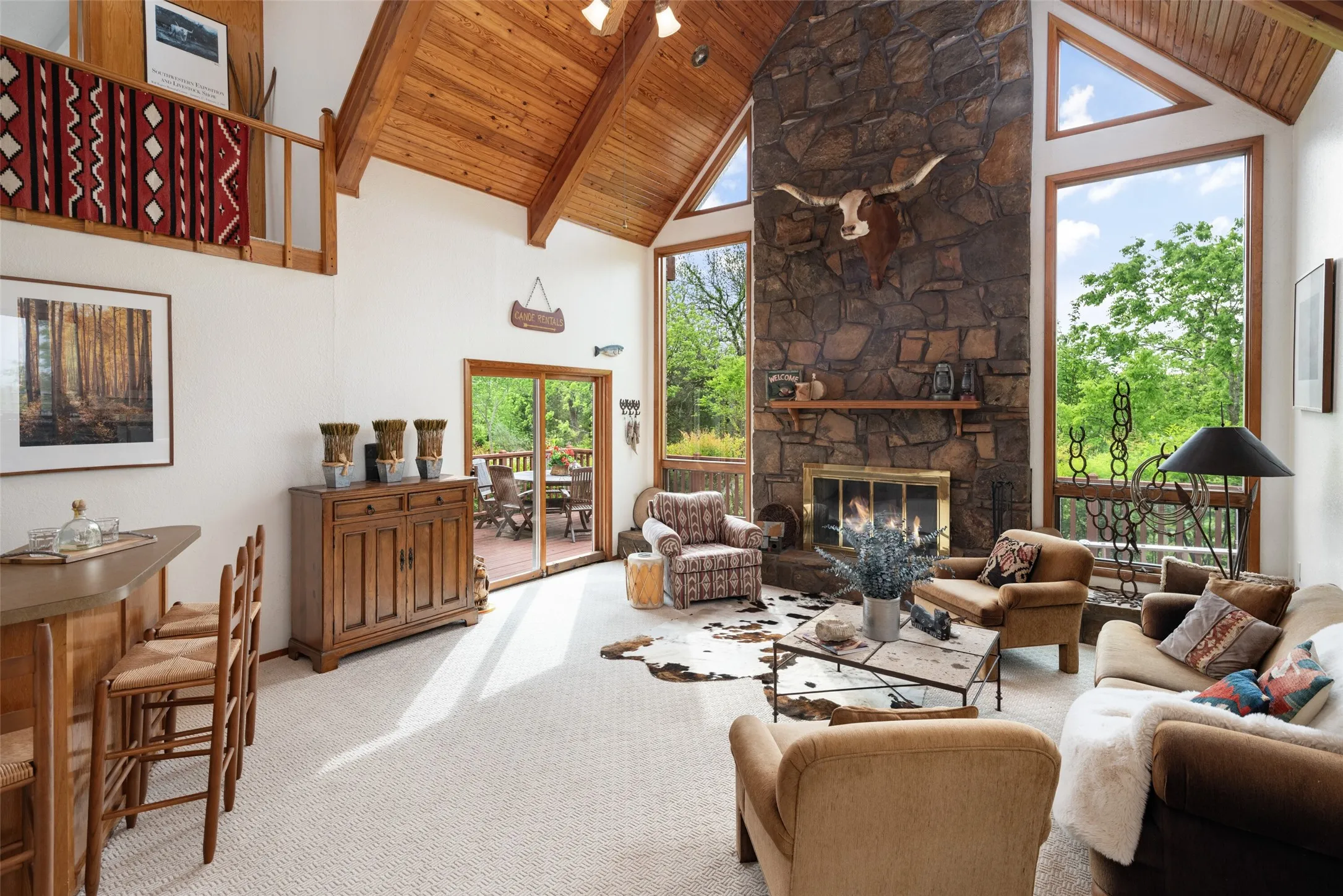 Vaulted wood-finish ceilings with exposed beams, stone-clad fireplace, large picture windows, and a sliding glass door to an outdoor deck
