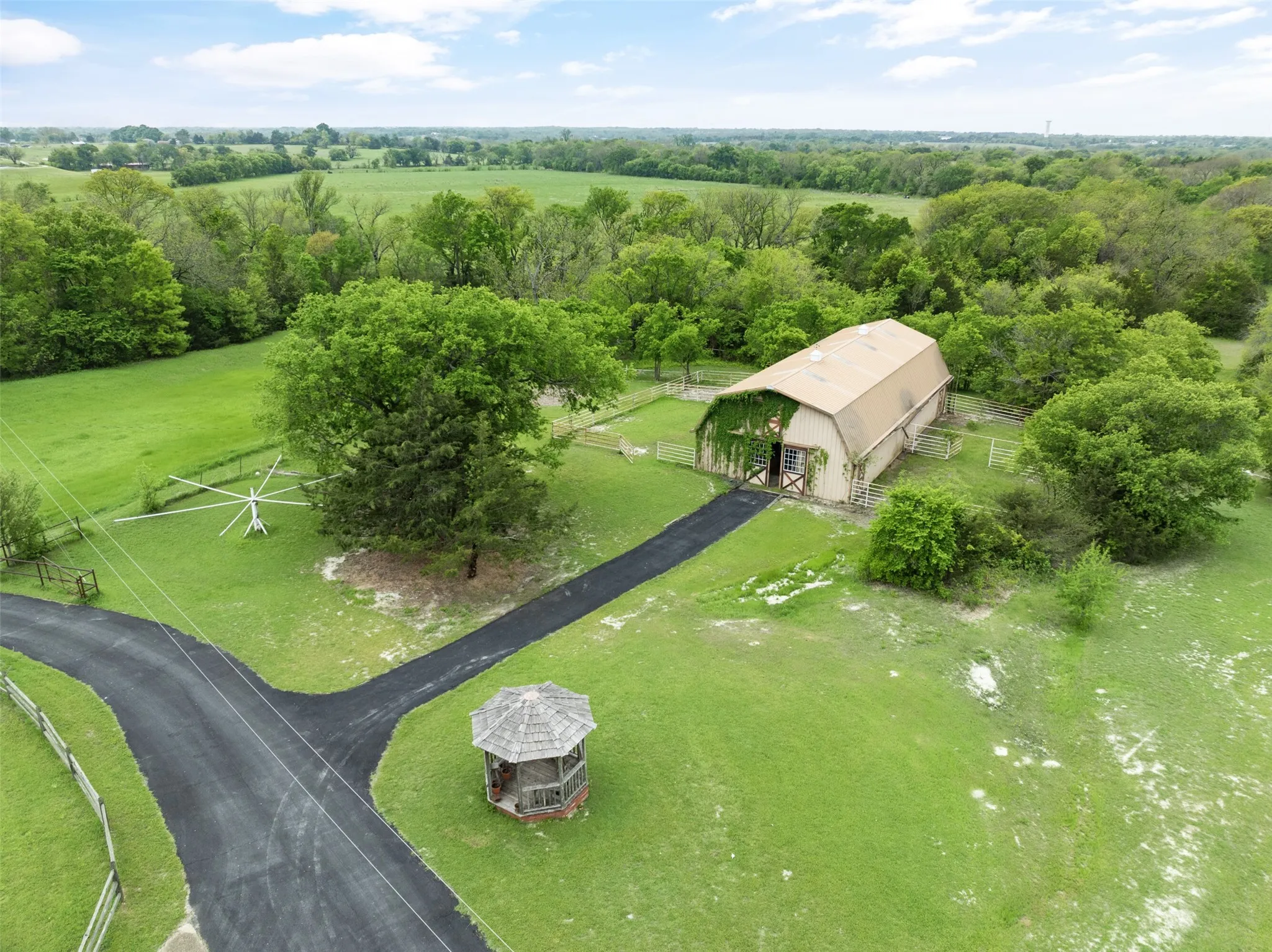 Expansive green acreage featuring a barn with a gambrel roof, mature trees, paved driveways, and a wooden gazebo