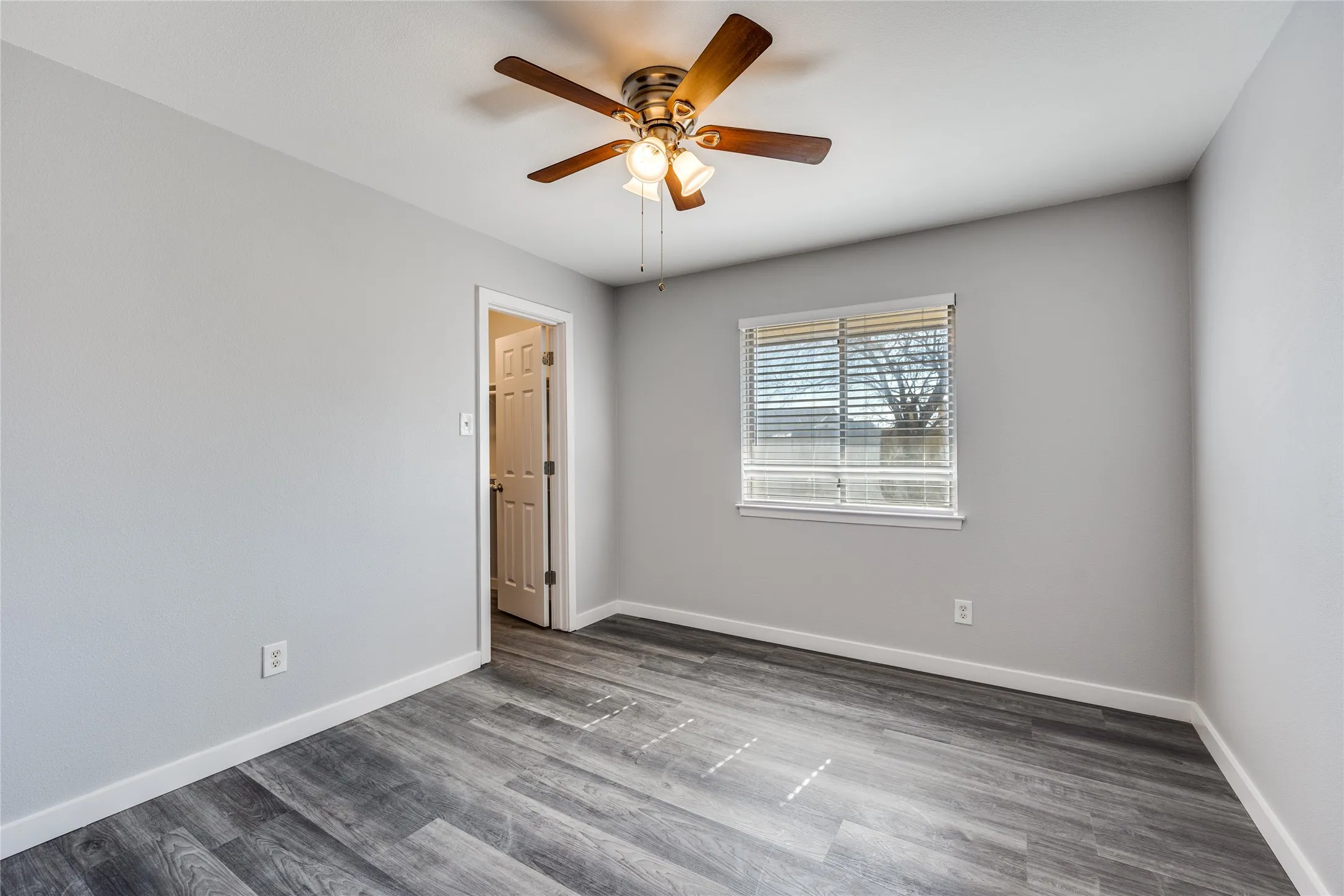 Light gray wall paint, wood-finish flooring, ceiling fan with integrated lighting, white window blinds, and a white interior door