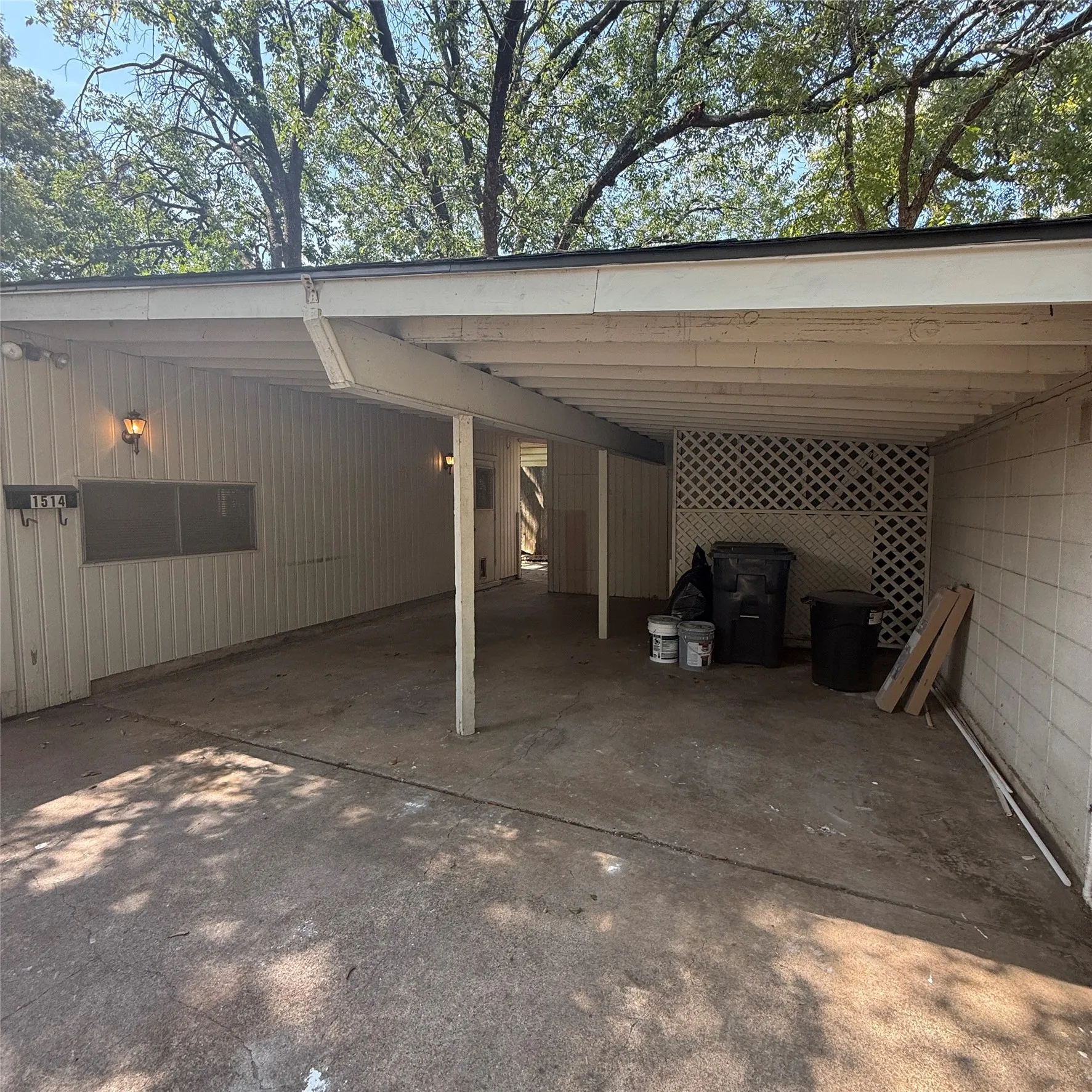 Carport structure with exposed rafters, concrete flooring, exterior wall lighting, and a lattice privacy screen