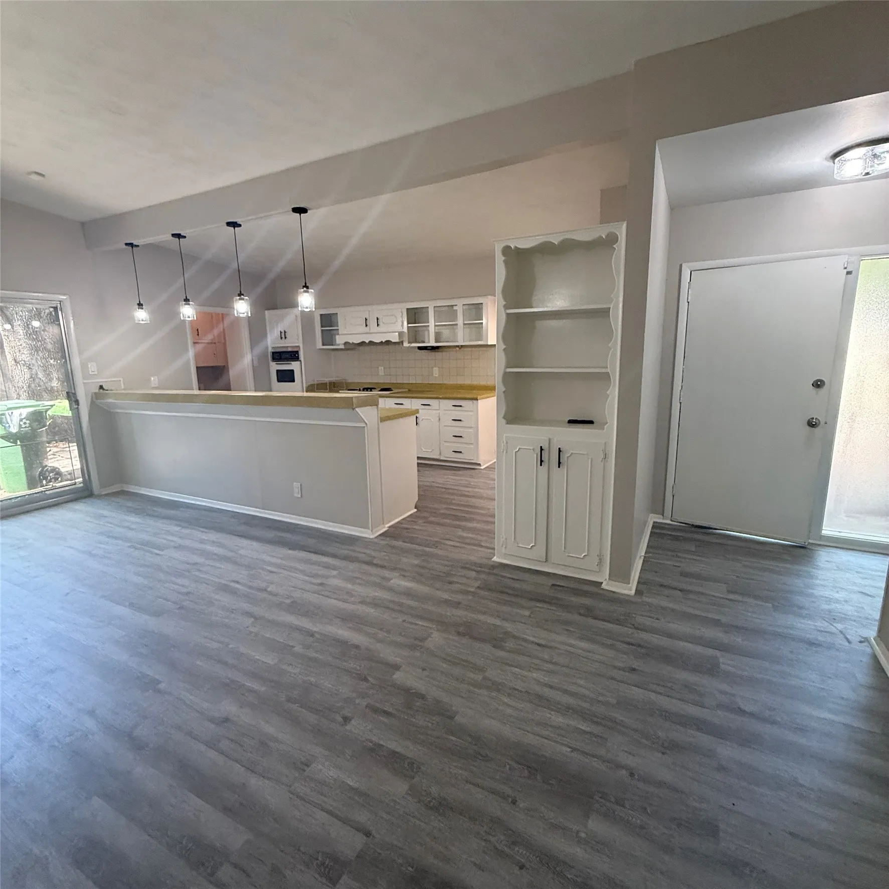 Open-concept living area with wood-finish flooring, built-in shelving, and a kitchen featuring white cabinetry, subway tile backsplash, and pendant lighting