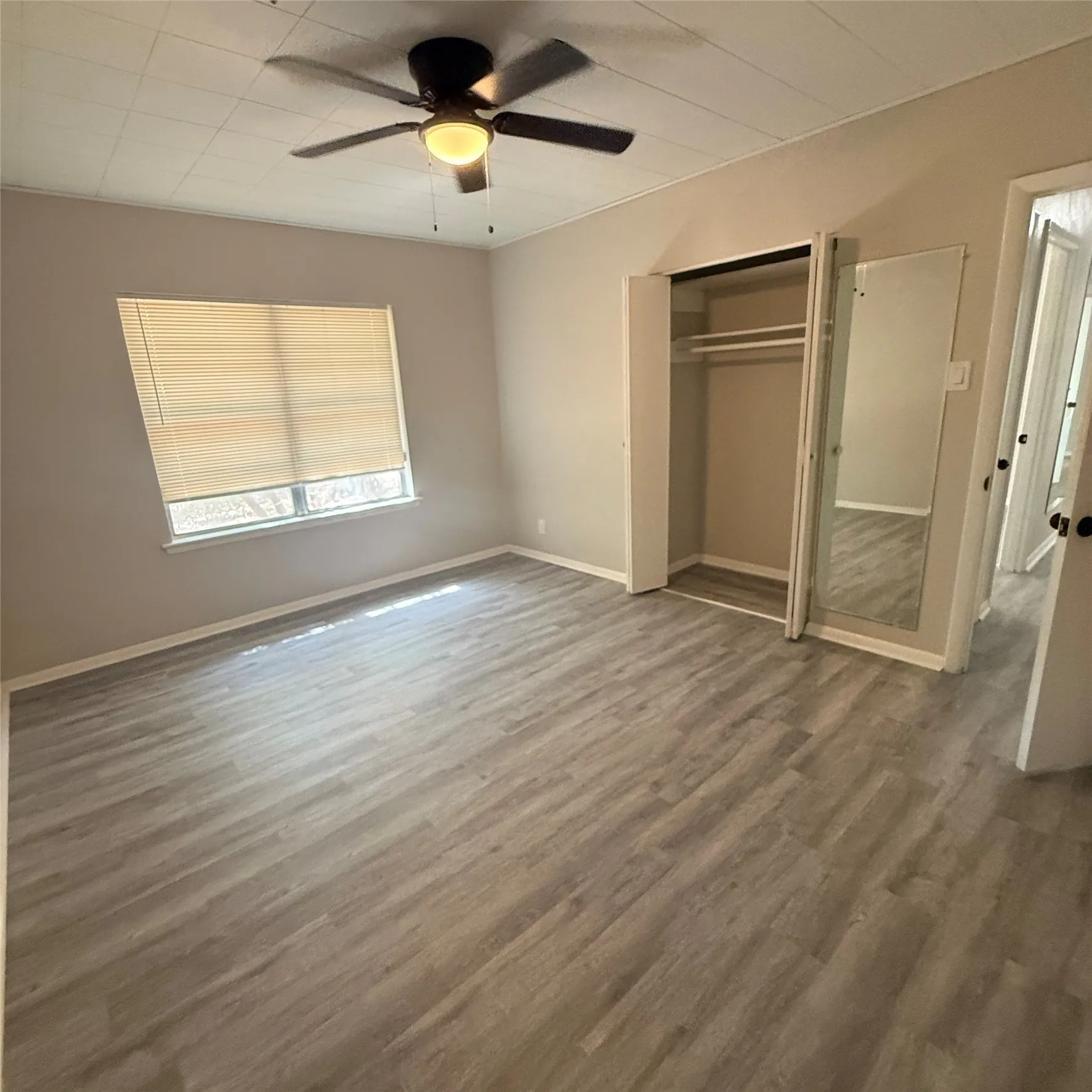 Spacious room featuring wood-finish flooring, a ceiling fan with integrated lighting, window with blinds, and a bi-fold closet with mirrored doors