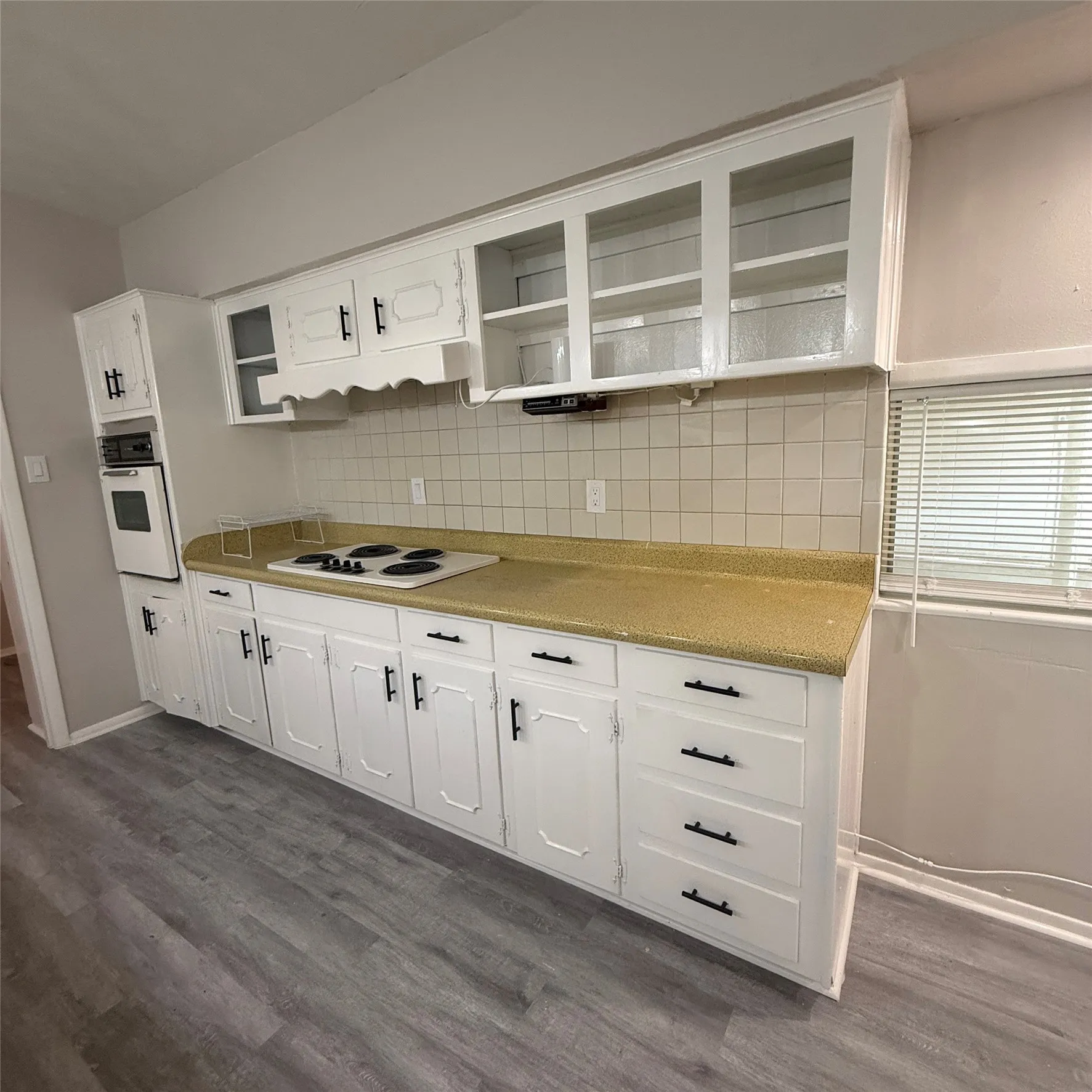 White cabinetry with dark hardware, built-in wall oven, cooktop, tan countertops, and a tiled backsplash