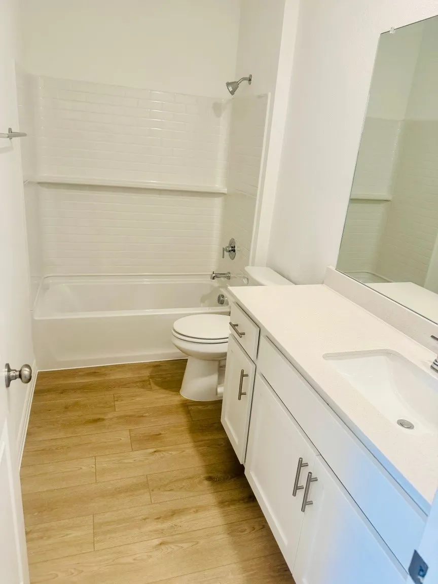 Wood-finish flooring, white subway tile shower surround, white vanity with integrated sink, chrome hardware, and a framed mirror