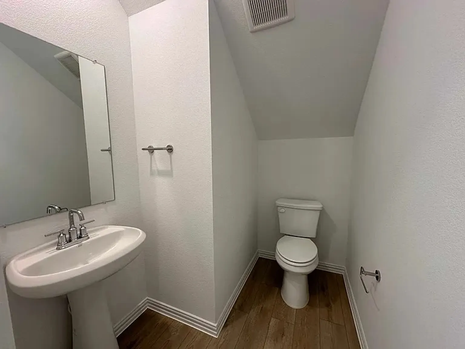 Pedestal sink with chrome faucet, wall-mounted mirror, wood-finish flooring, and white toilet