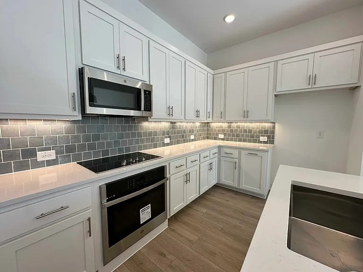 Kitchen featuring white shaker-style cabinetry, stainless steel appliances, a tile backsplash, light-toned countertops, and wood-finish flooring