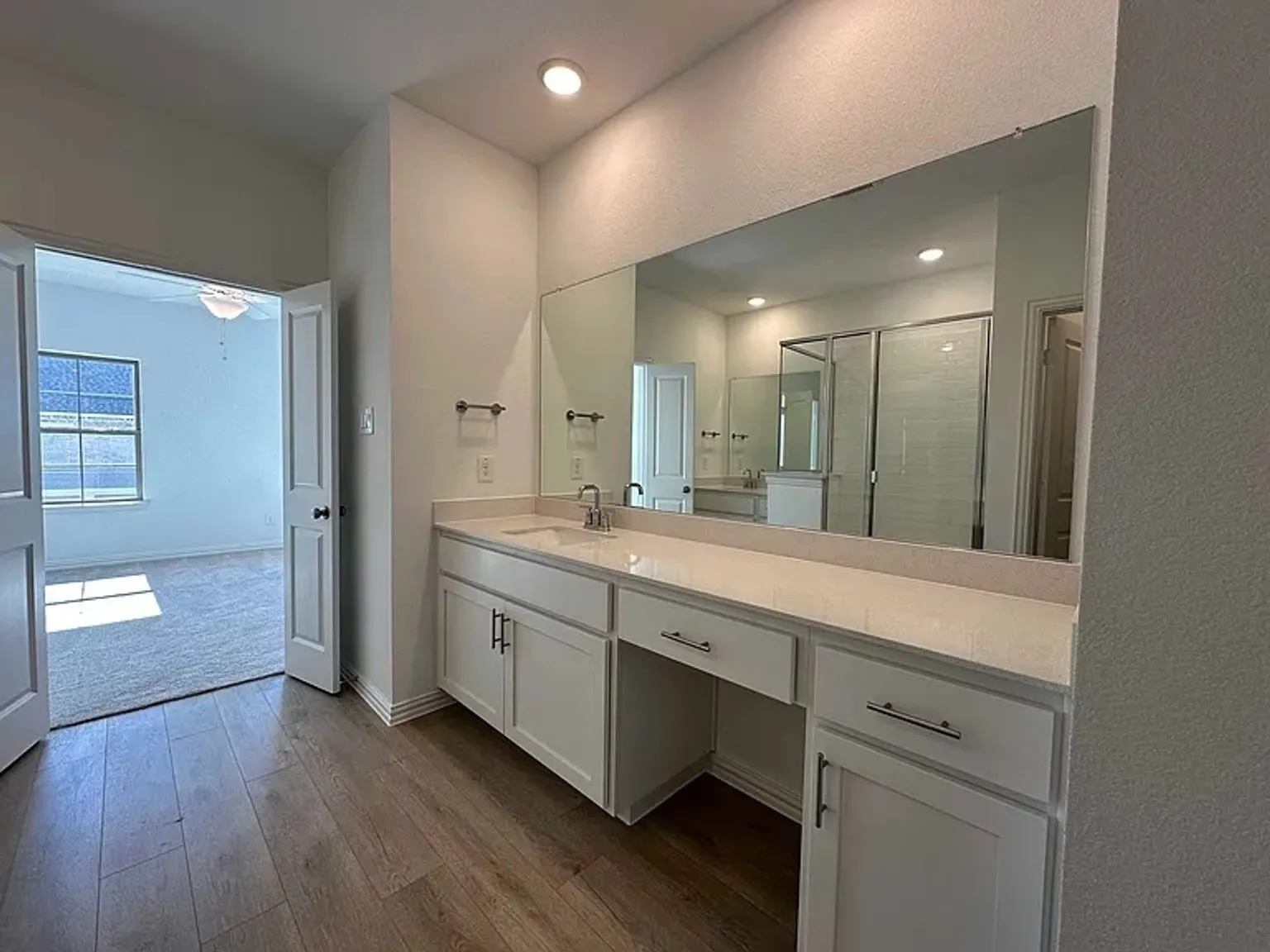 Wood-finish flooring leading to a bathroom featuring a white vanity with an integrated sink, stone countertop, and a large wall-mounted mirror