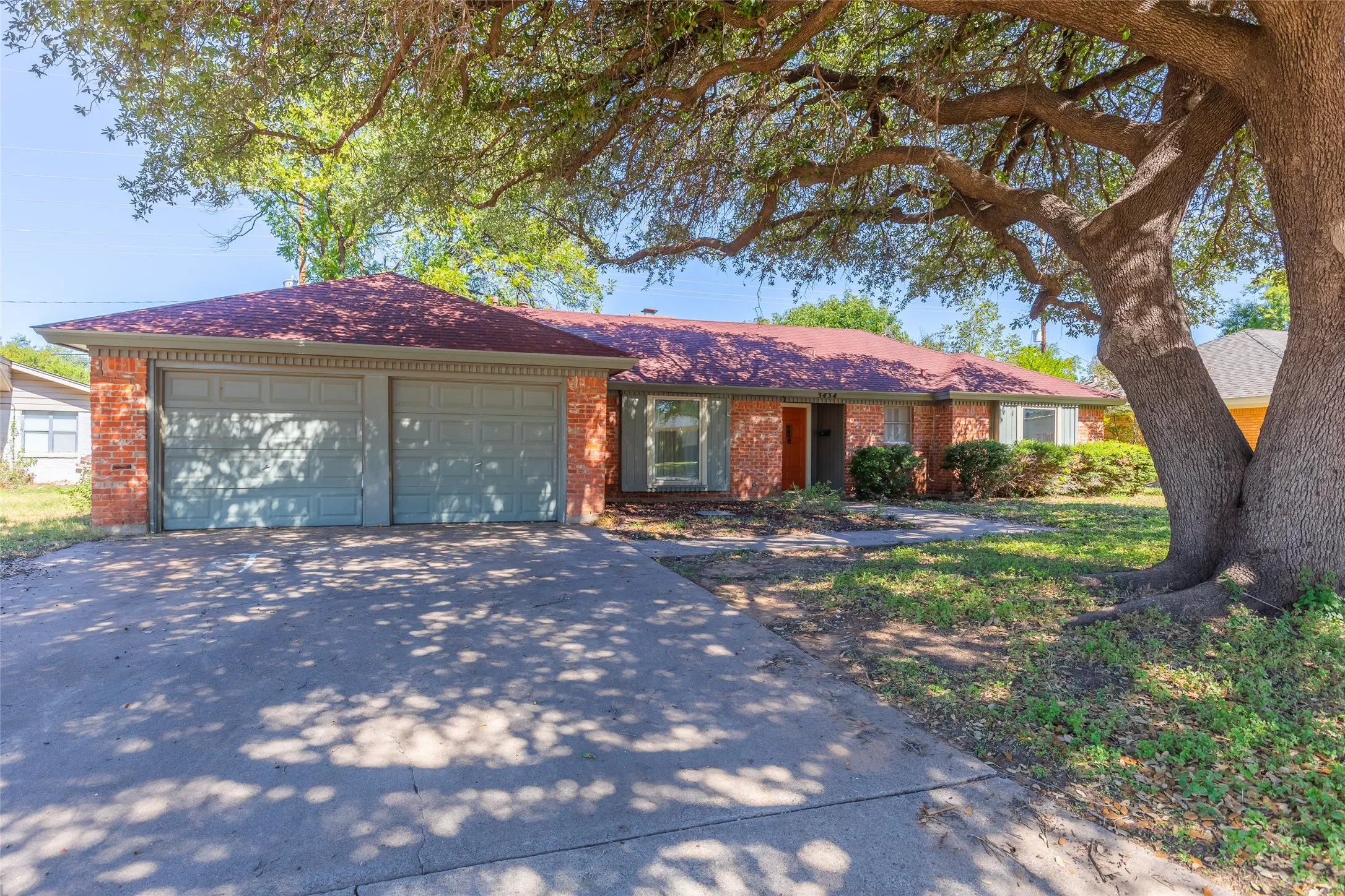 Brick exterior with a two-car garage, red shingle roof, and mature trees providing natural shade