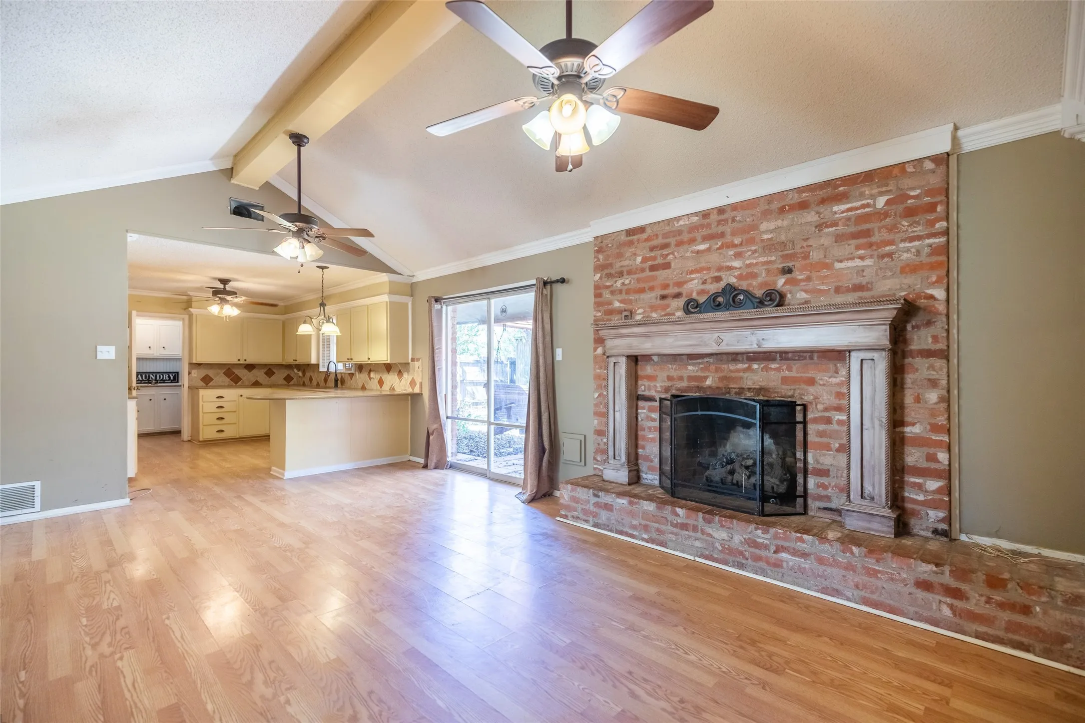 Vaulted ceiling with exposed beams, wood-finish flooring, brick fireplace with wood mantel, sliding glass door, and recessed kitchen with built-in cabinetry