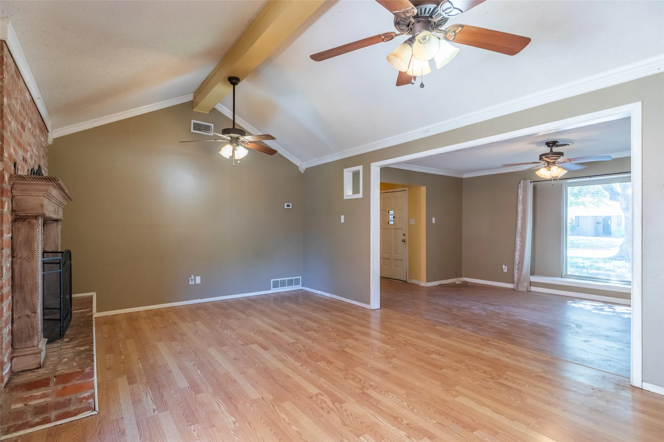 Vaulted ceiling with exposed wood beam, wood-finish flooring, brick fireplace with wood mantel, multiple ceiling fans, and a broad window