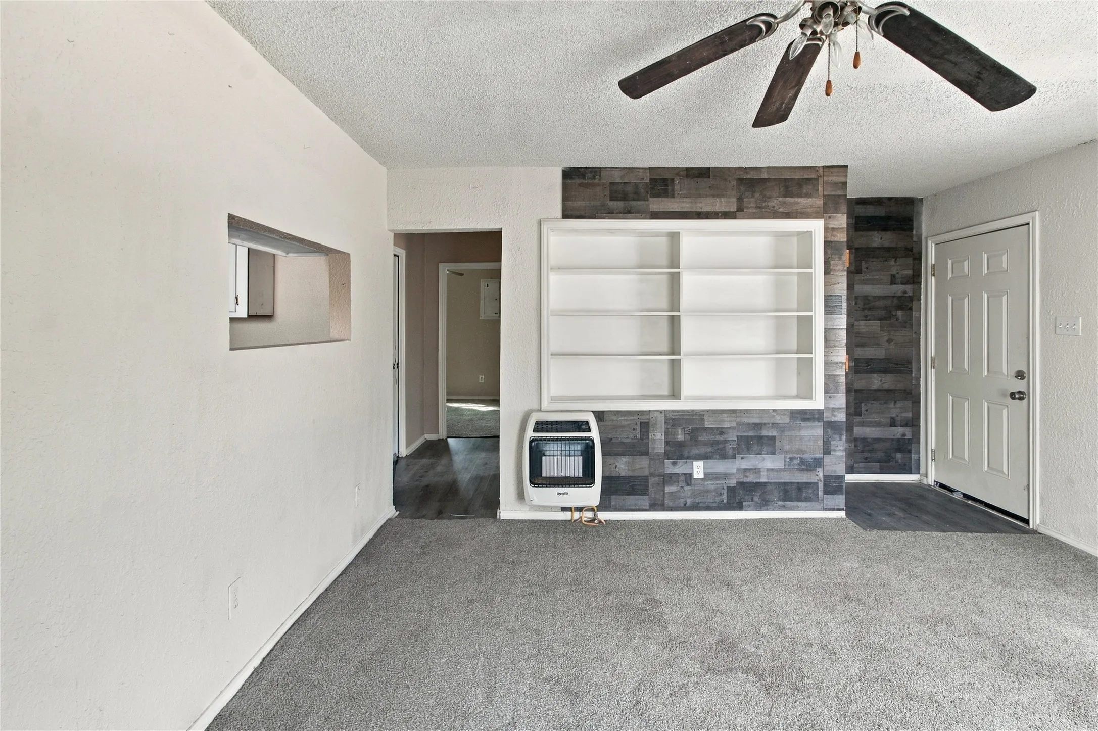 Gray carpet flooring, white built-in shelving, wood-finish accent wall, ceiling fan with light fixture, and pass-through window