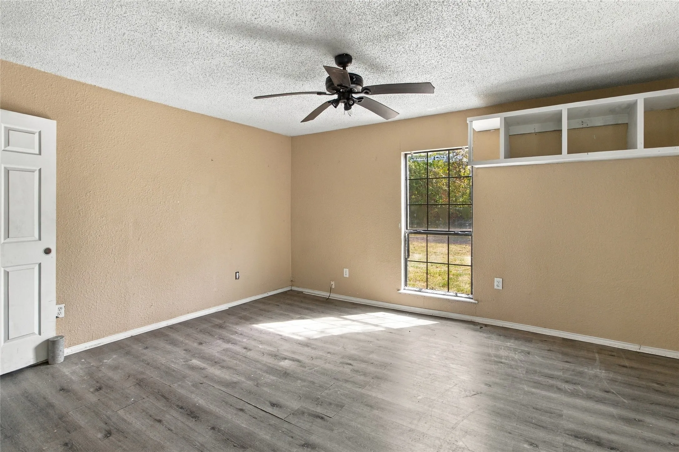 Spacious room with wood-finish flooring, ceiling fan with remote control, large windows providing natural light, built-in cabinetry with glass-front upper shelving, and neutral wall finishes
