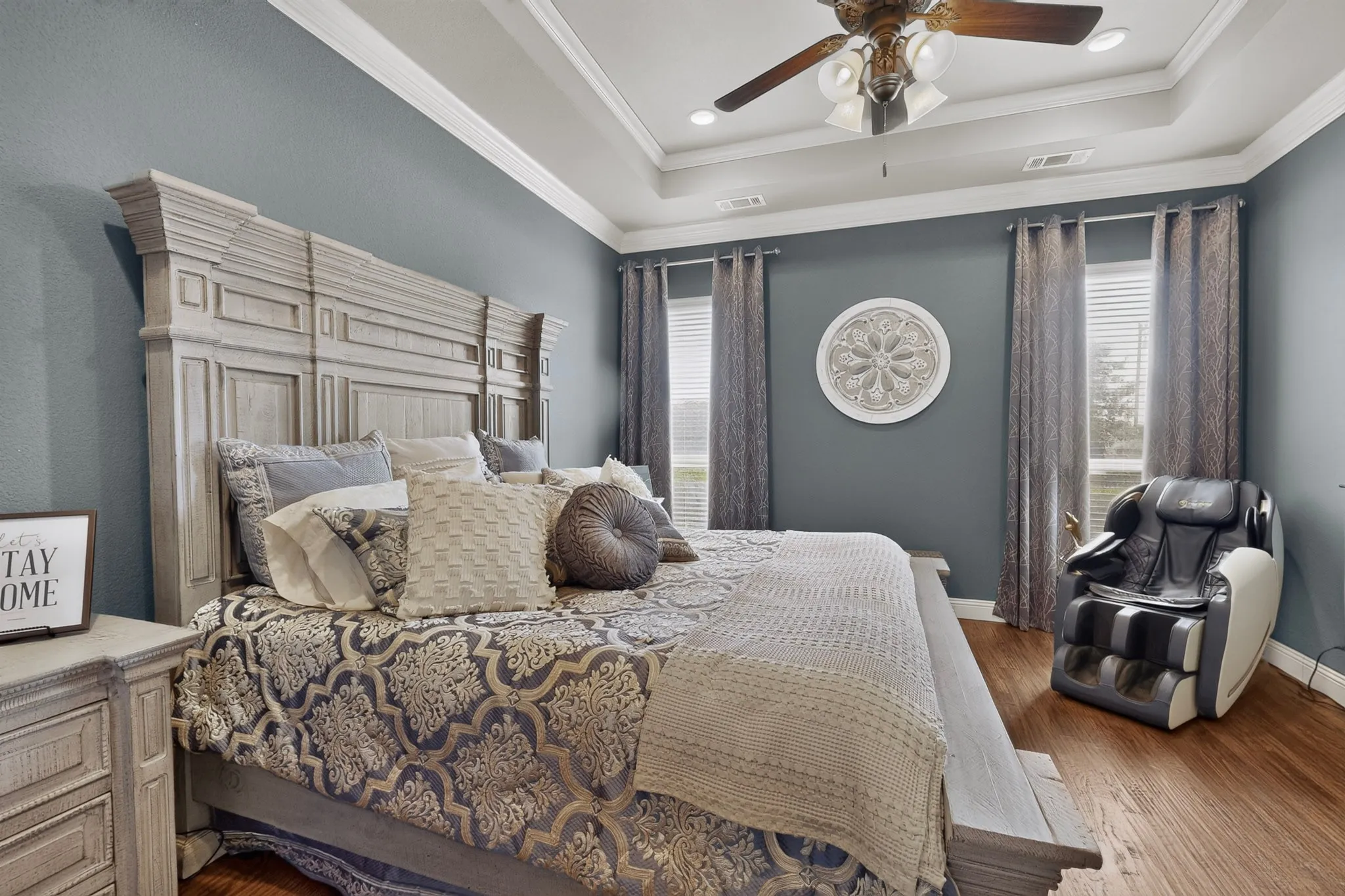 Master bedroom featuring soft blue walls, a decorative tray ceiling with ceiling fan, hardwood flooring, and large windows with coordinating drapery