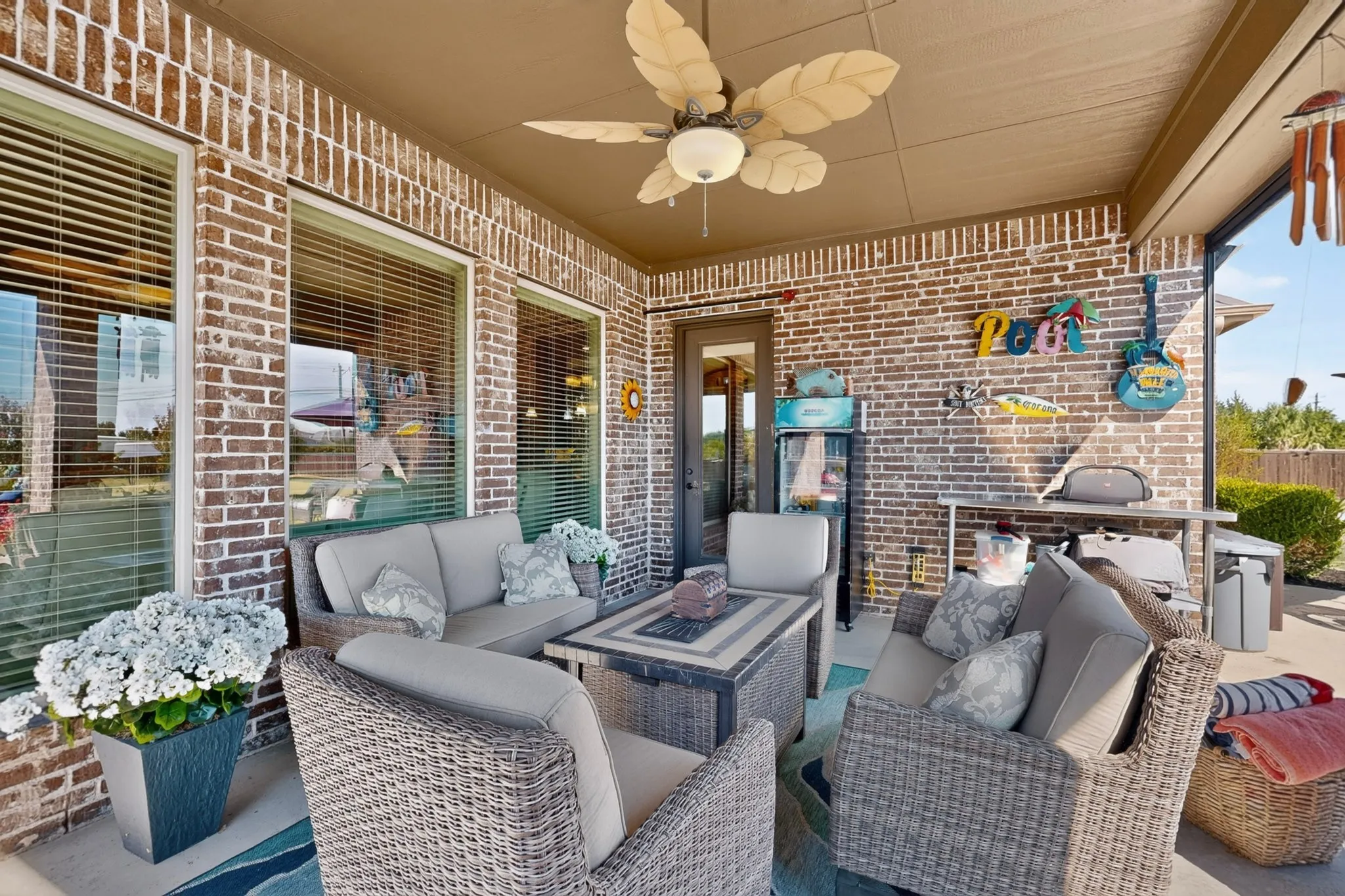 Covered porch with brick exterior featuring comfortable wicker seating, a fire table with metal frame, ceiling fan with integrated lighting, and large windows allowing natural light throughout the space