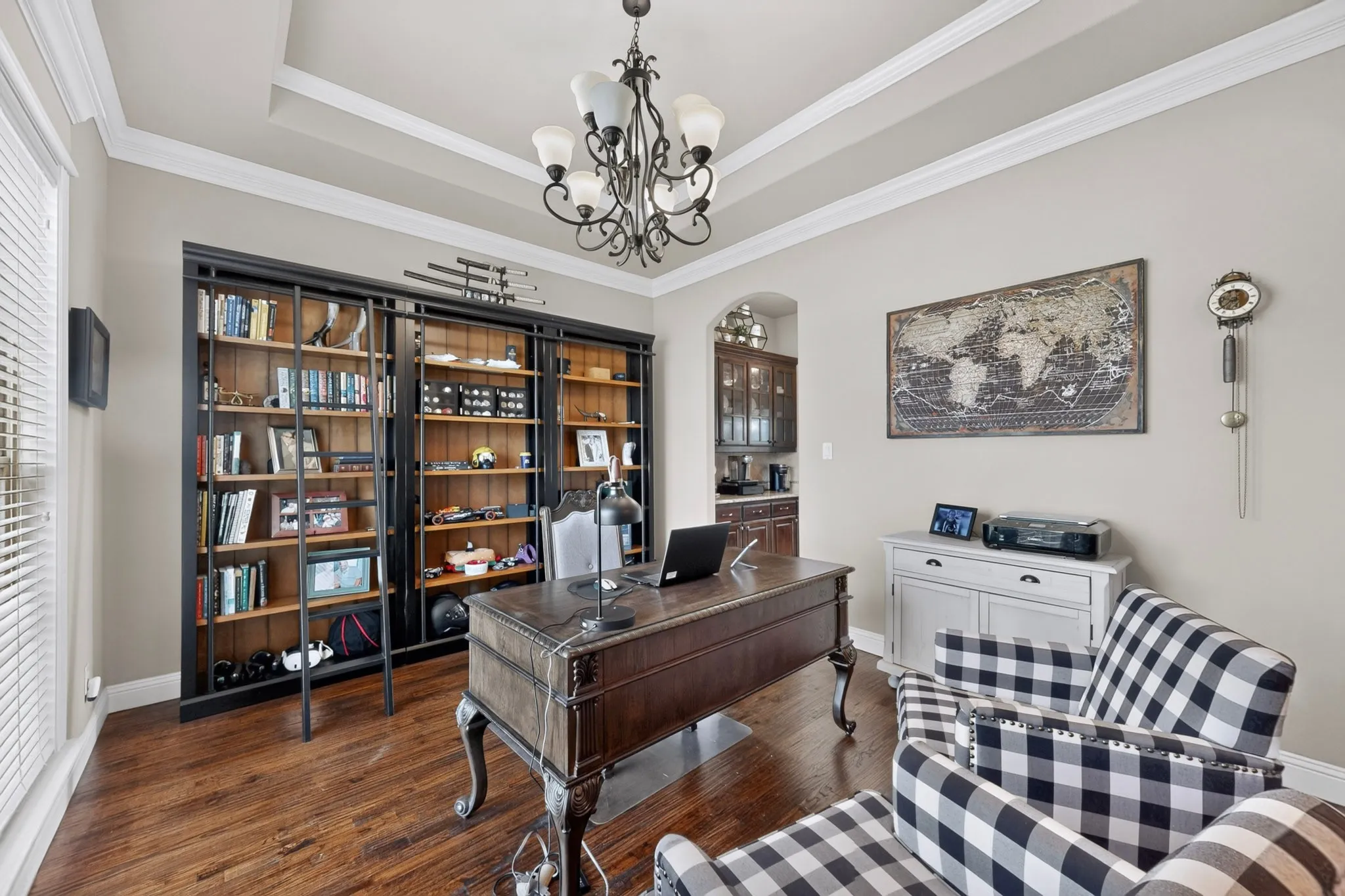 Sophisticated home office featuring rich wood-finish flooring, extensive built-in cabinetry with glass-front display shelving, a traditional desk with decorative turned legs, coffered ceiling with crown molding, and a coordinating upholstered accent chair