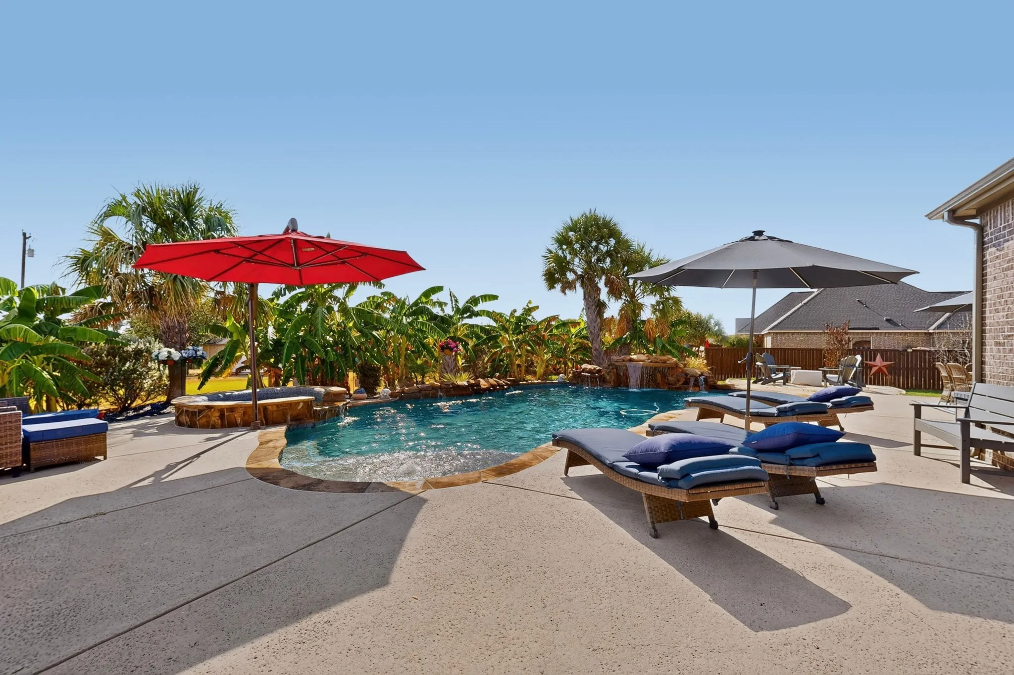 Curved swimming pool with a stone waterfall feature, expansive concrete patio, and lush tropical landscaping
