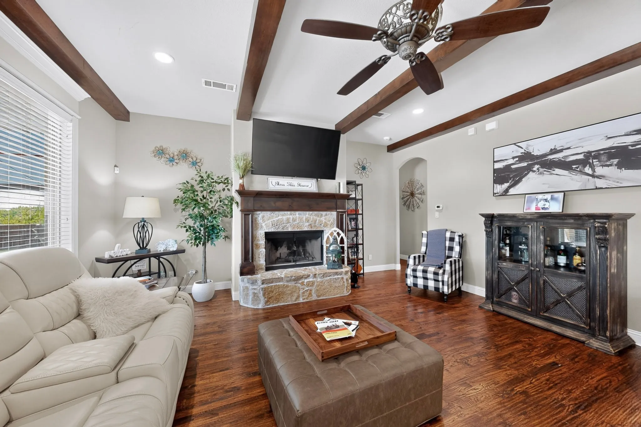 Spacious living area featuring exposed wood beam ceiling details, a wood-finish plank floor throughout, and a fireplace with stone surround