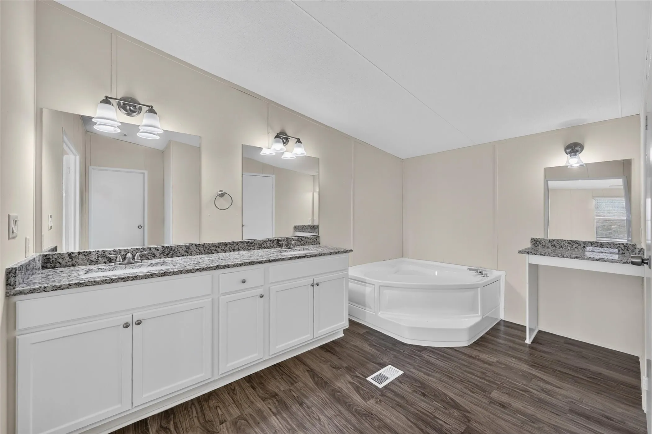 Wood-finish flooring, white cabinetry, granite countertops, dual vanity mirrors, and a corner soaking tub