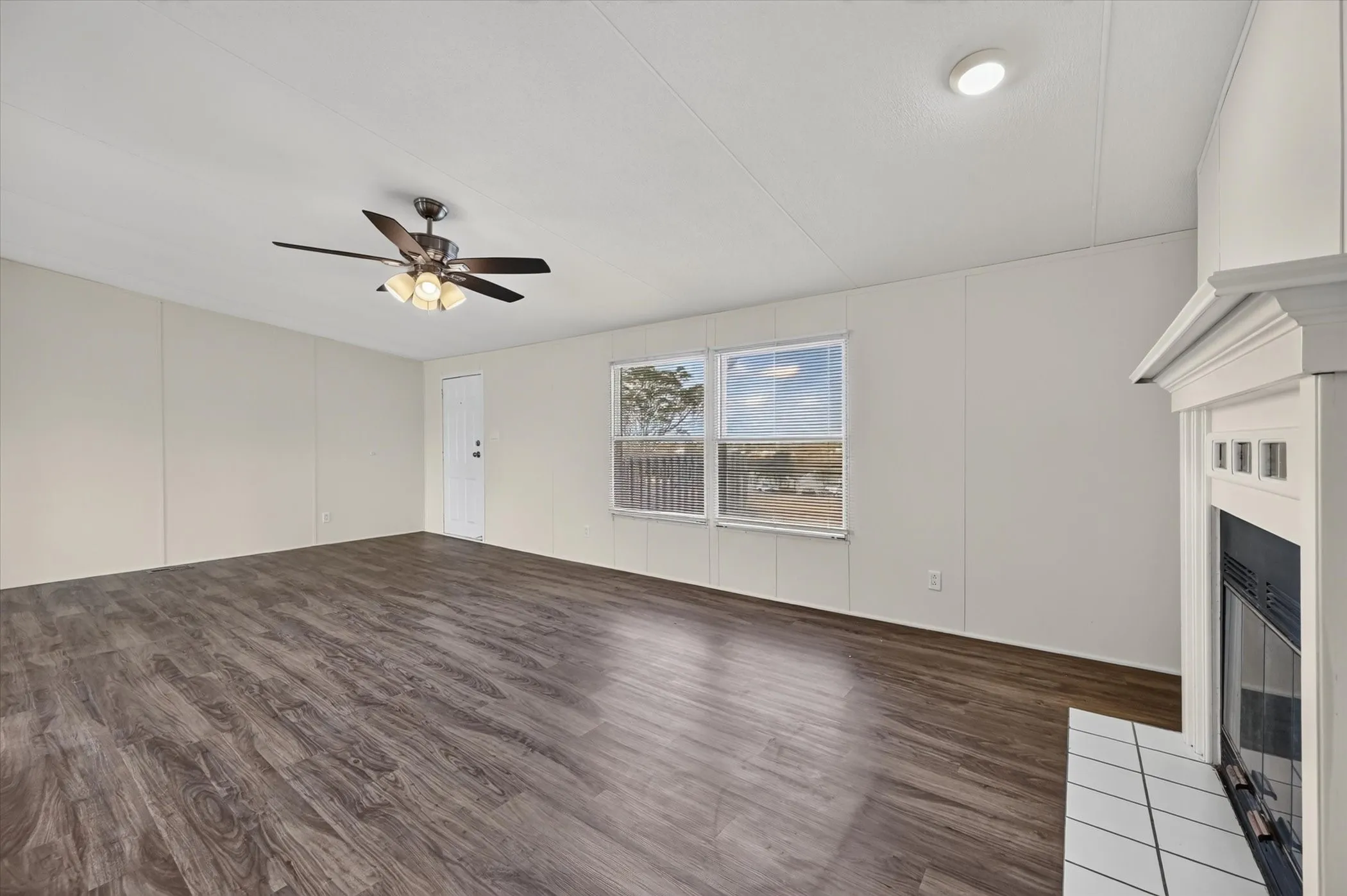 Spacious interior featuring wood-finish flooring, a ceiling fan with integrated lighting, multiple windows, a recessed ceiling light, and a fireplace with a white mantel and tiled hearth