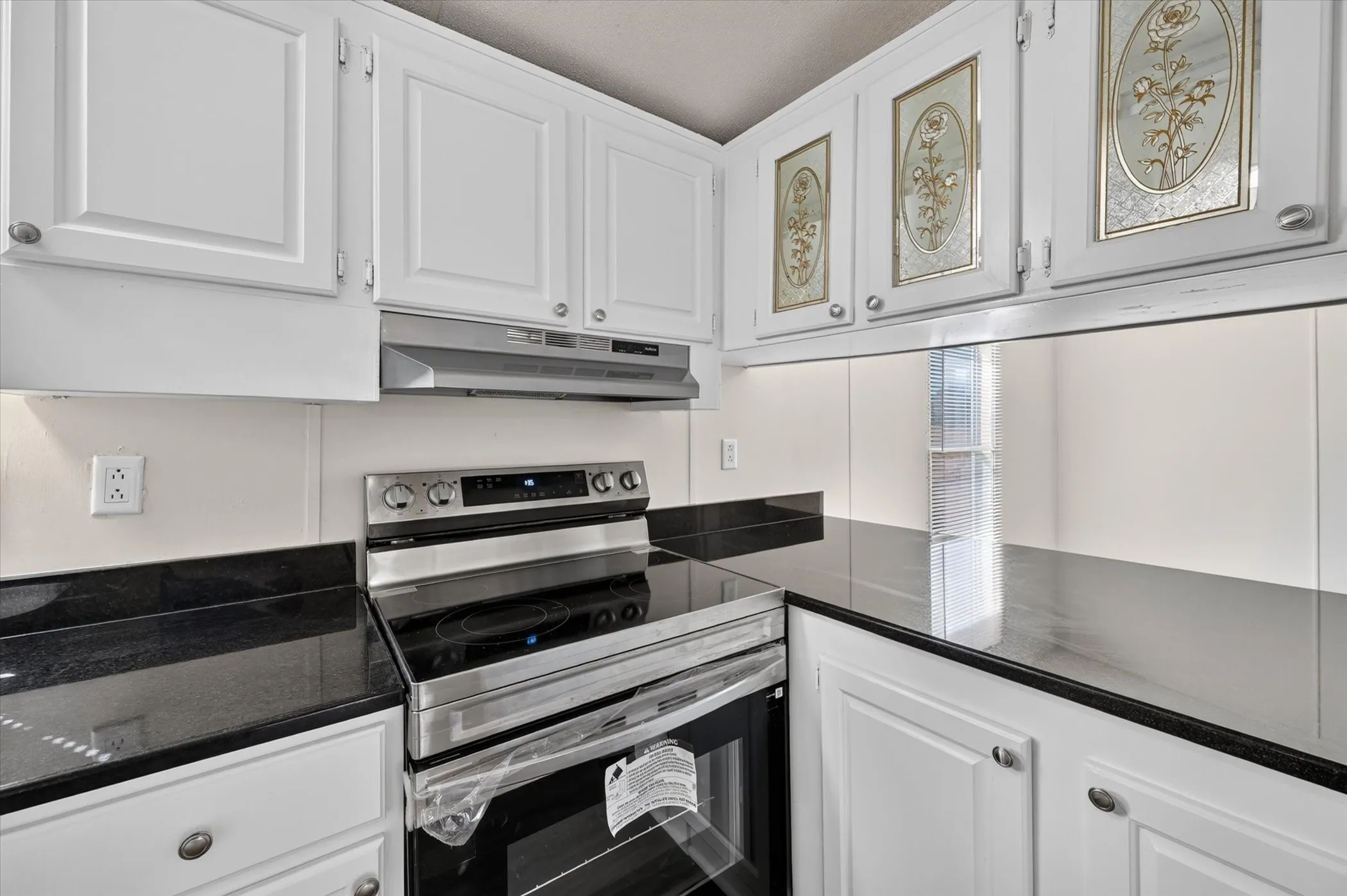 White cabinetry with decorative glass inserts, black countertops, stainless steel range, and matching range hood