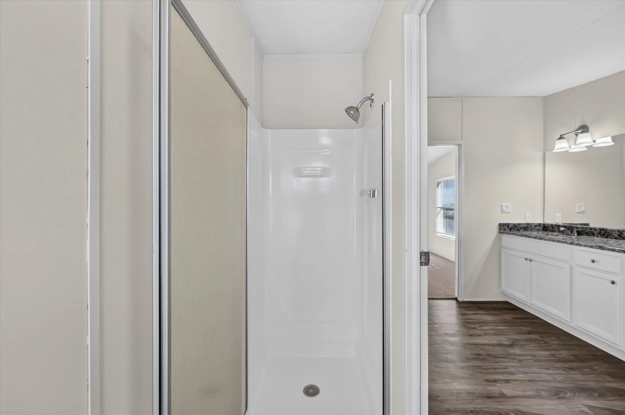 Shower with a sliding glass enclosure, a white vanity with dual sinks, a dark countertop, and wood-finish flooring