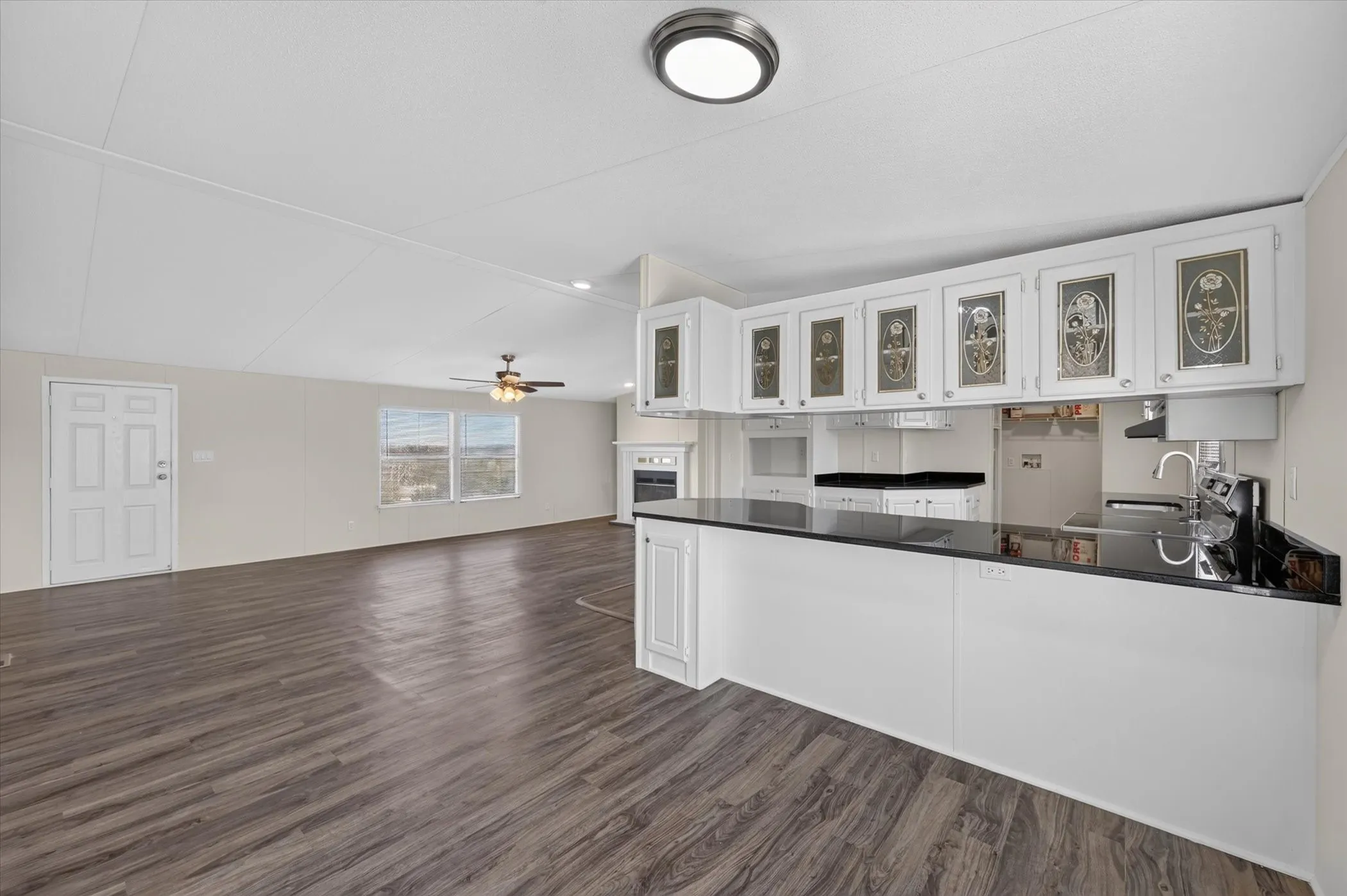 Open-concept living space with wood-finish flooring, white cabinetry with decorative glass inserts, a black countertop, and a ceiling fan