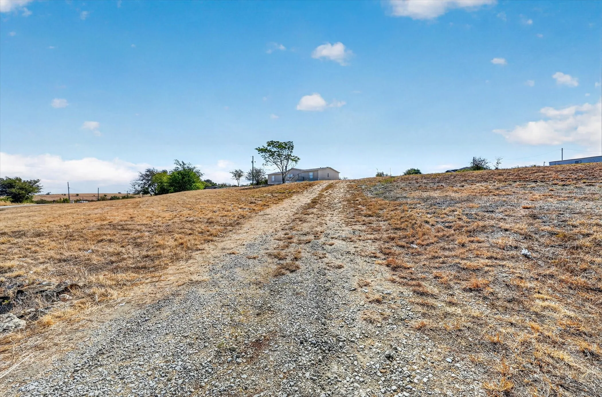 Expansive undeveloped land featuring a gravel driveway and a distant residential structure