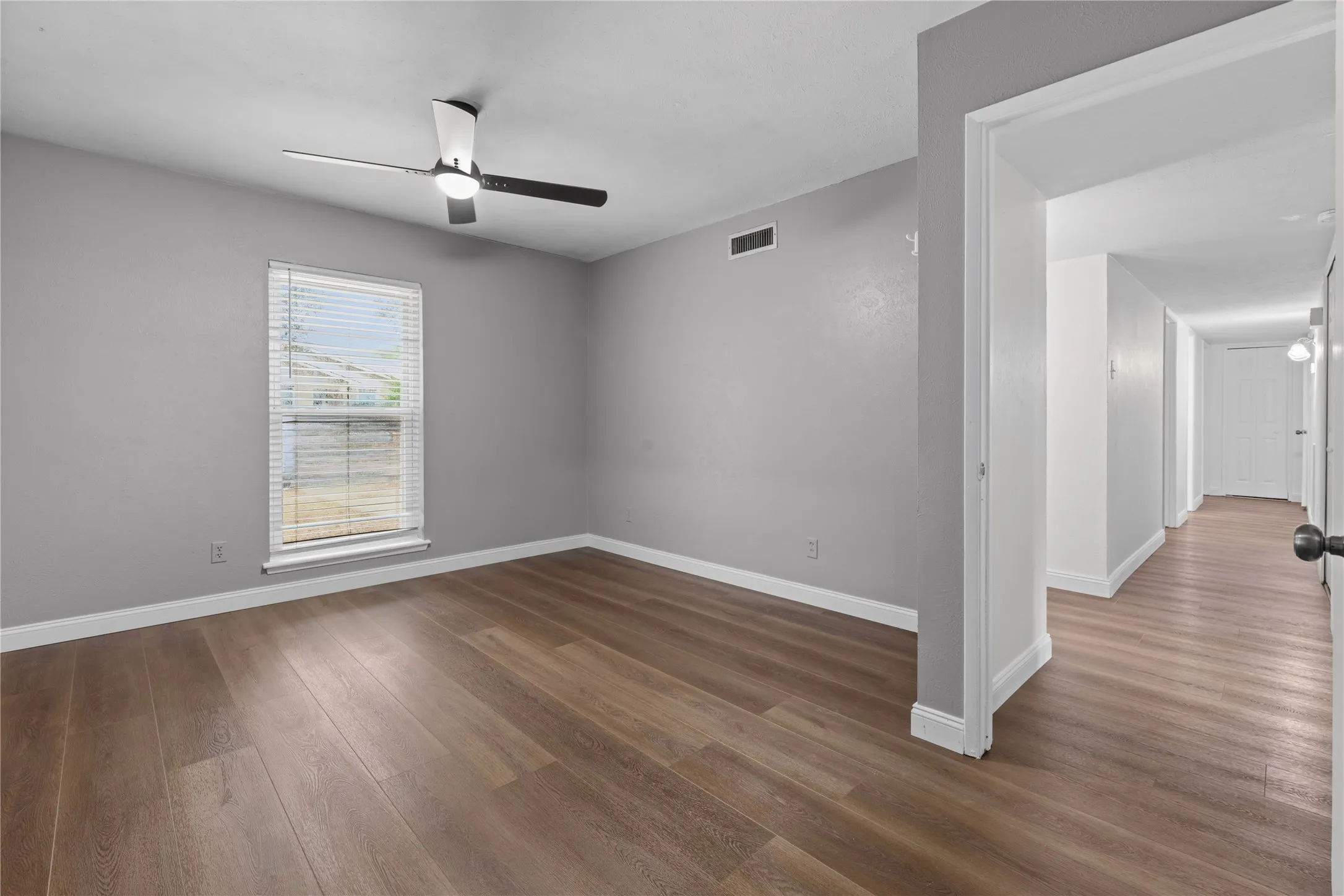 Wood-finish flooring throughout, light gray wall paint, a ceiling fan with integrated lighting, and a window with blinds