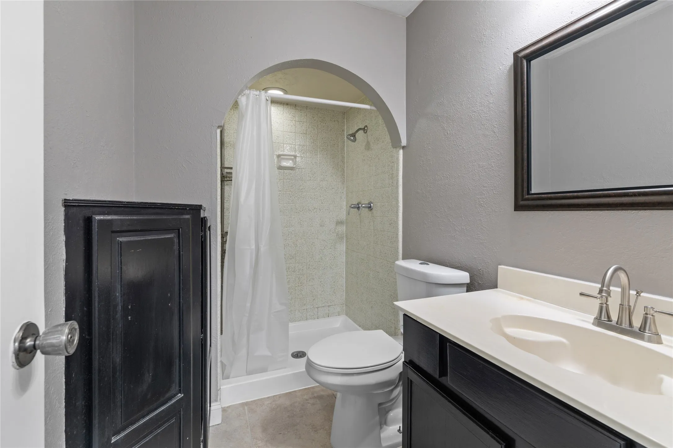 Bathroom featuring an arched shower enclosure with tile surround, a vanity with a white countertop and dark cabinetry, and a framed mirror