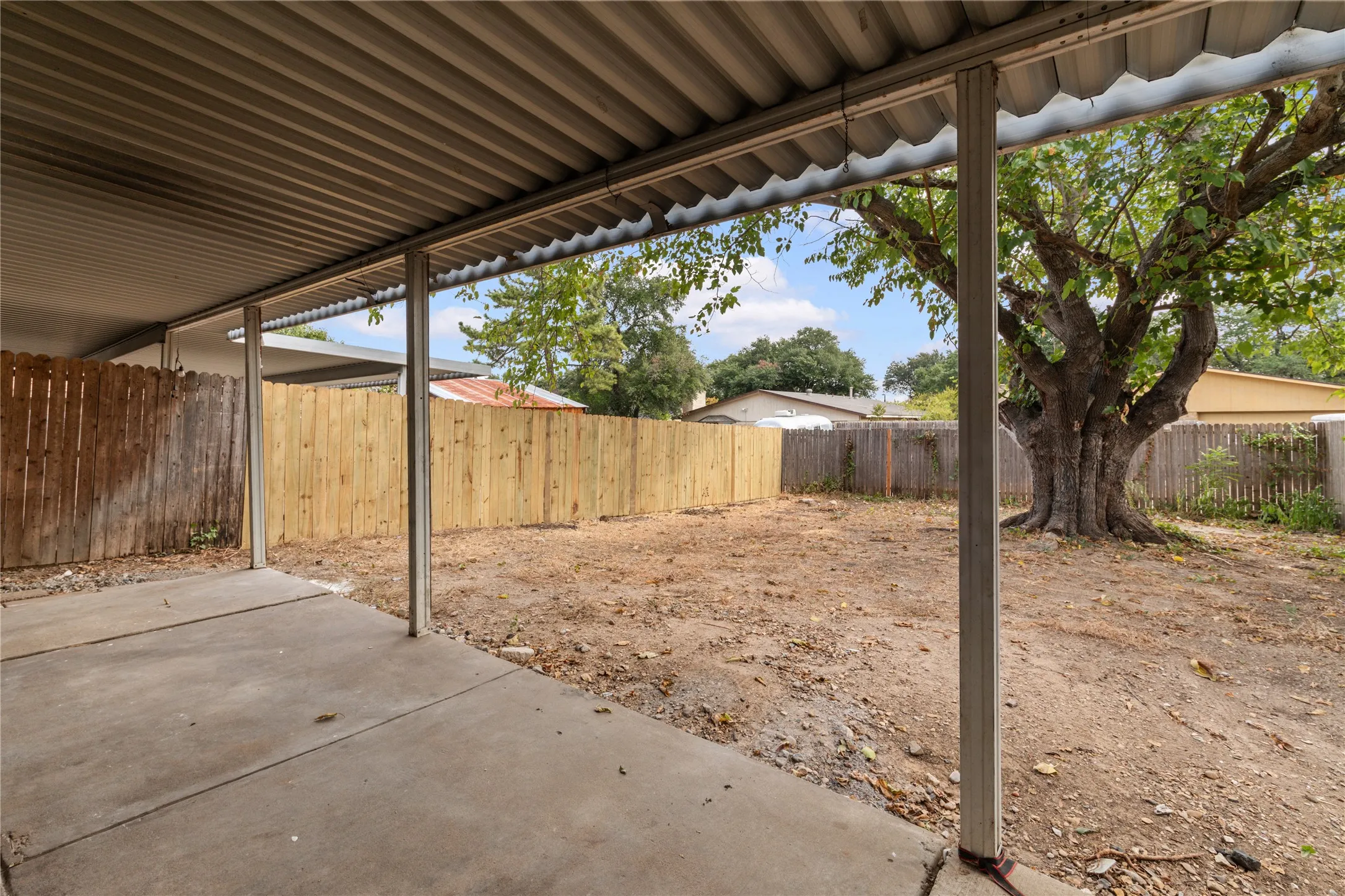 Covered patio with concrete flooring and metal beam construction overlooking a spacious backyard featuring wooden perimeter fencing and mature shade trees
