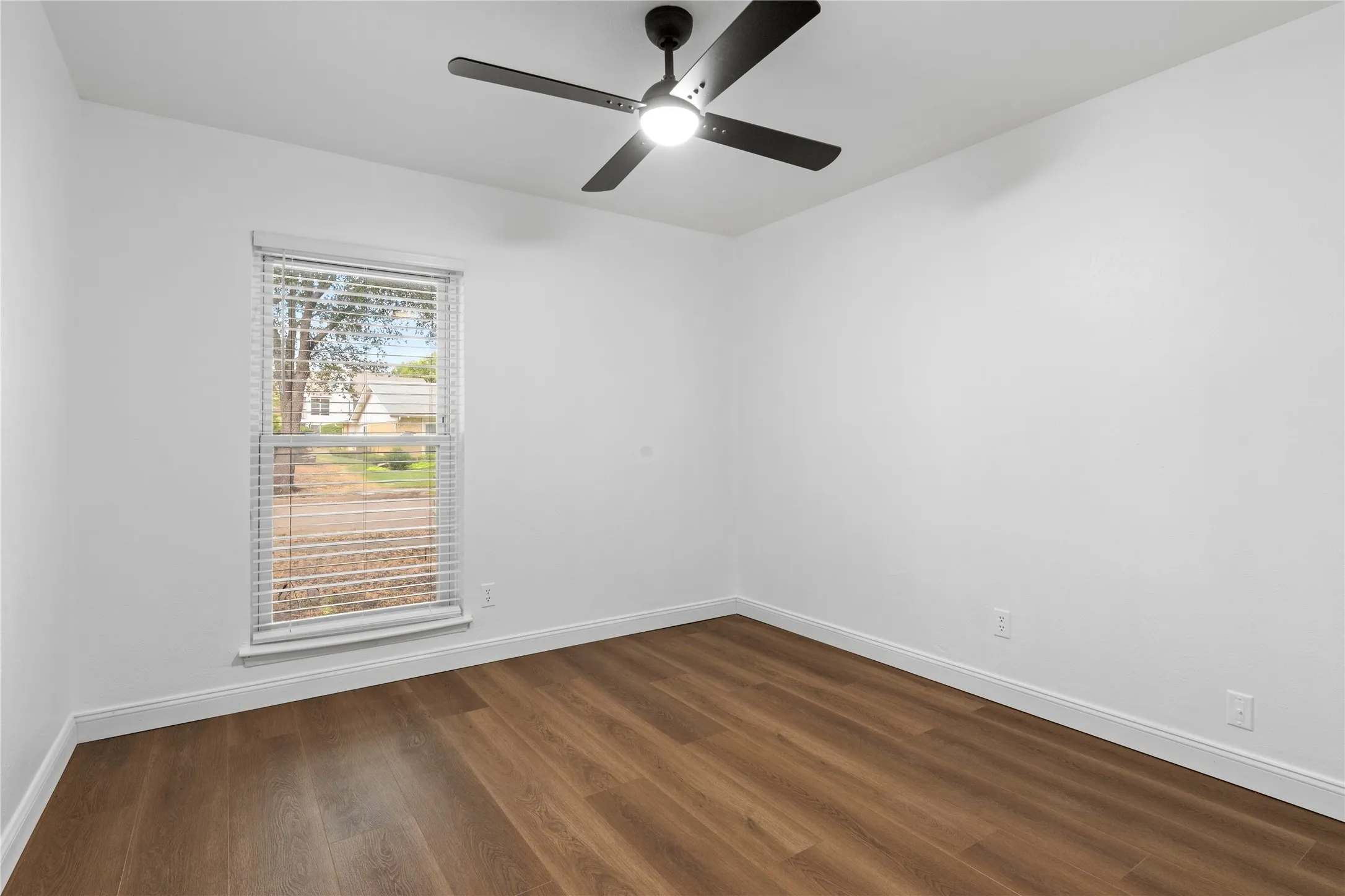 Clean white walls with wood-finish flooring, featuring a window with blinds, and a ceiling fan with integrated lighting