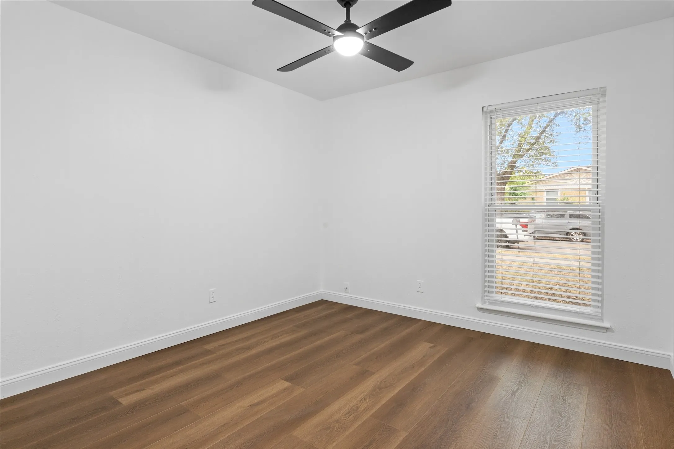 Bright corner bedroom featuring hardwood flooring, a ceiling fan with integrated lighting, and abundant natural light from a large picture window with horizontal blinds